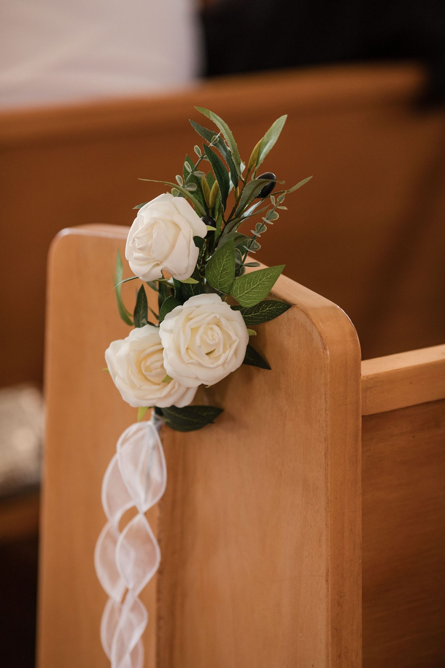 White roses with greenery tied with ribbon decorate a wooden church pew for a wedding ceremony.