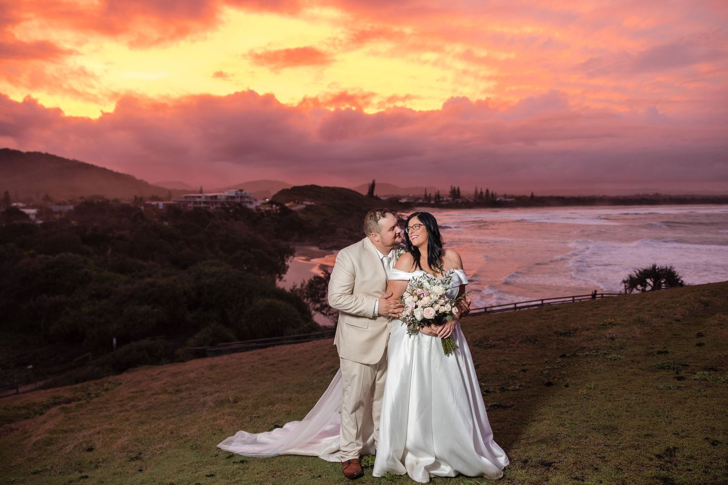 Bride and groom embrace at sunset on a hilltop overlooking a coastal bay with a dramatic pink and orange sky.