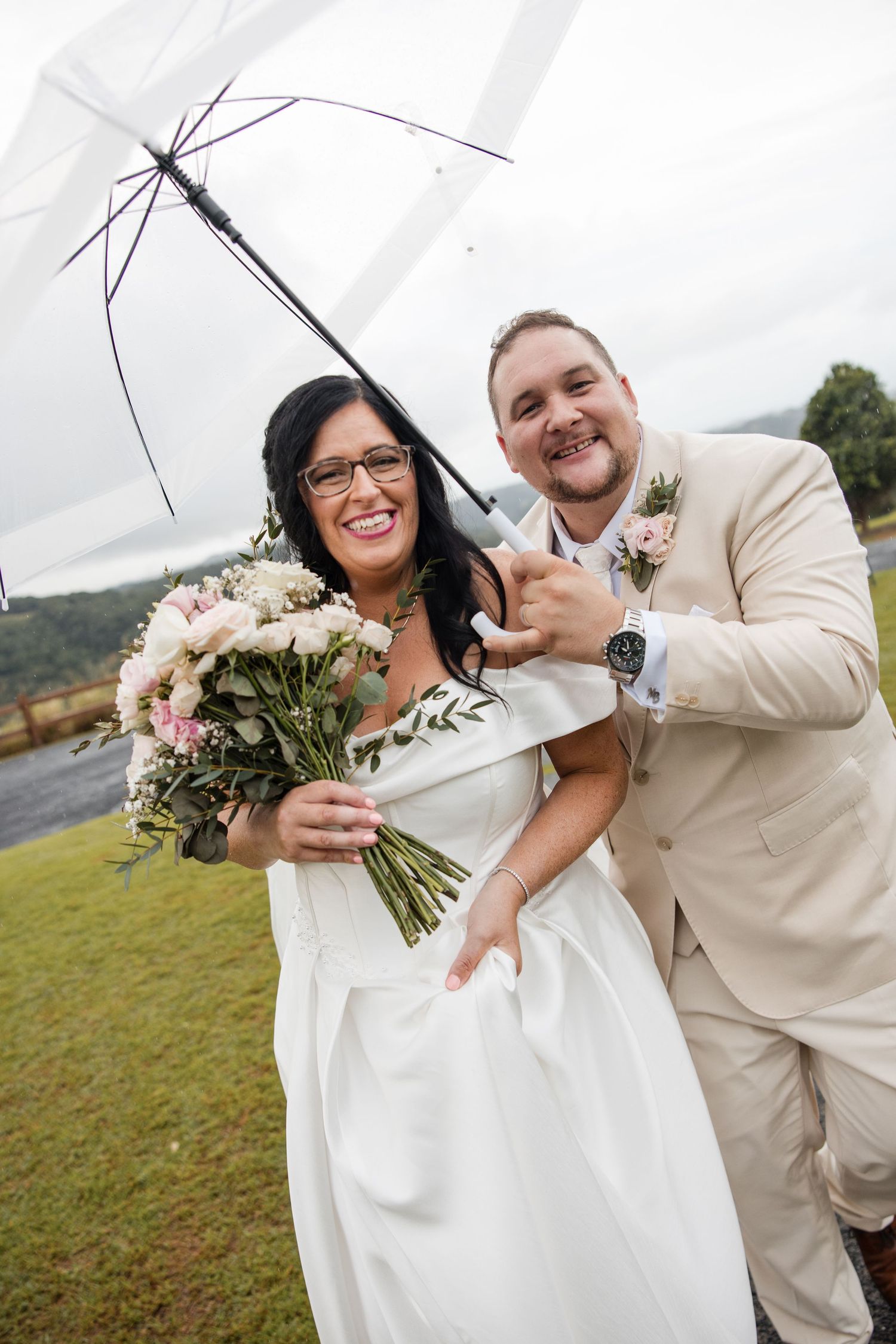 Bride and groom smiling under a clear umbrella on their wedding day, bride holding a pink bouquet in a white gown.