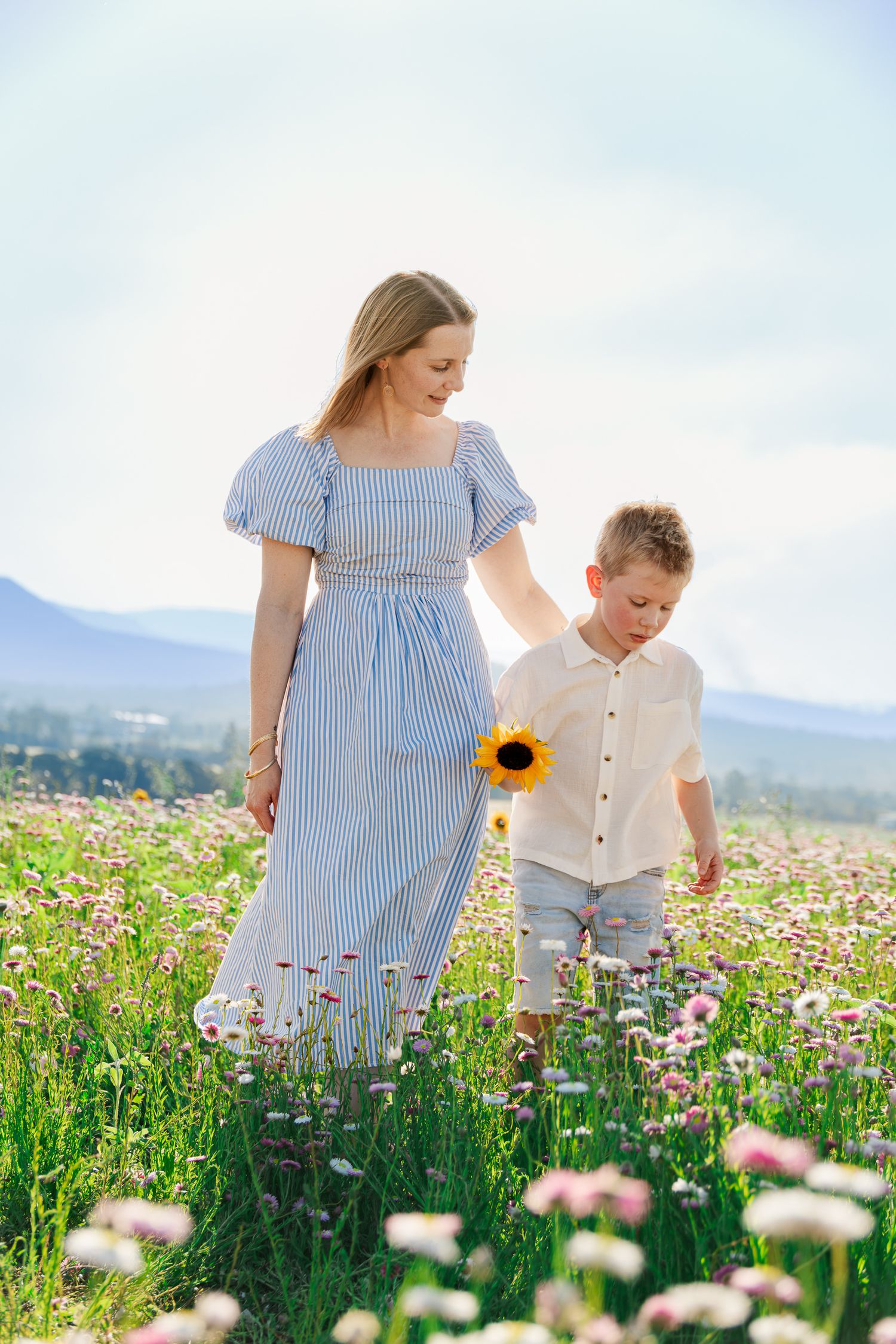 A mother in a blue striped dress walks with her young son holding a sunflower in a blooming wildflower field.