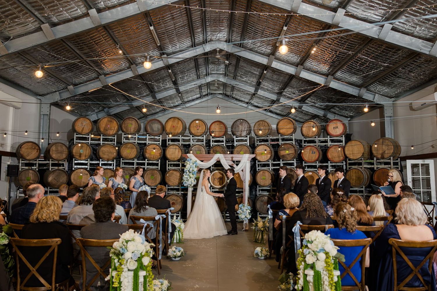 Wedding ceremony in a rustic barn venue with wine barrel backdrop, floral arrangements, and guests seated in wooden chairs.