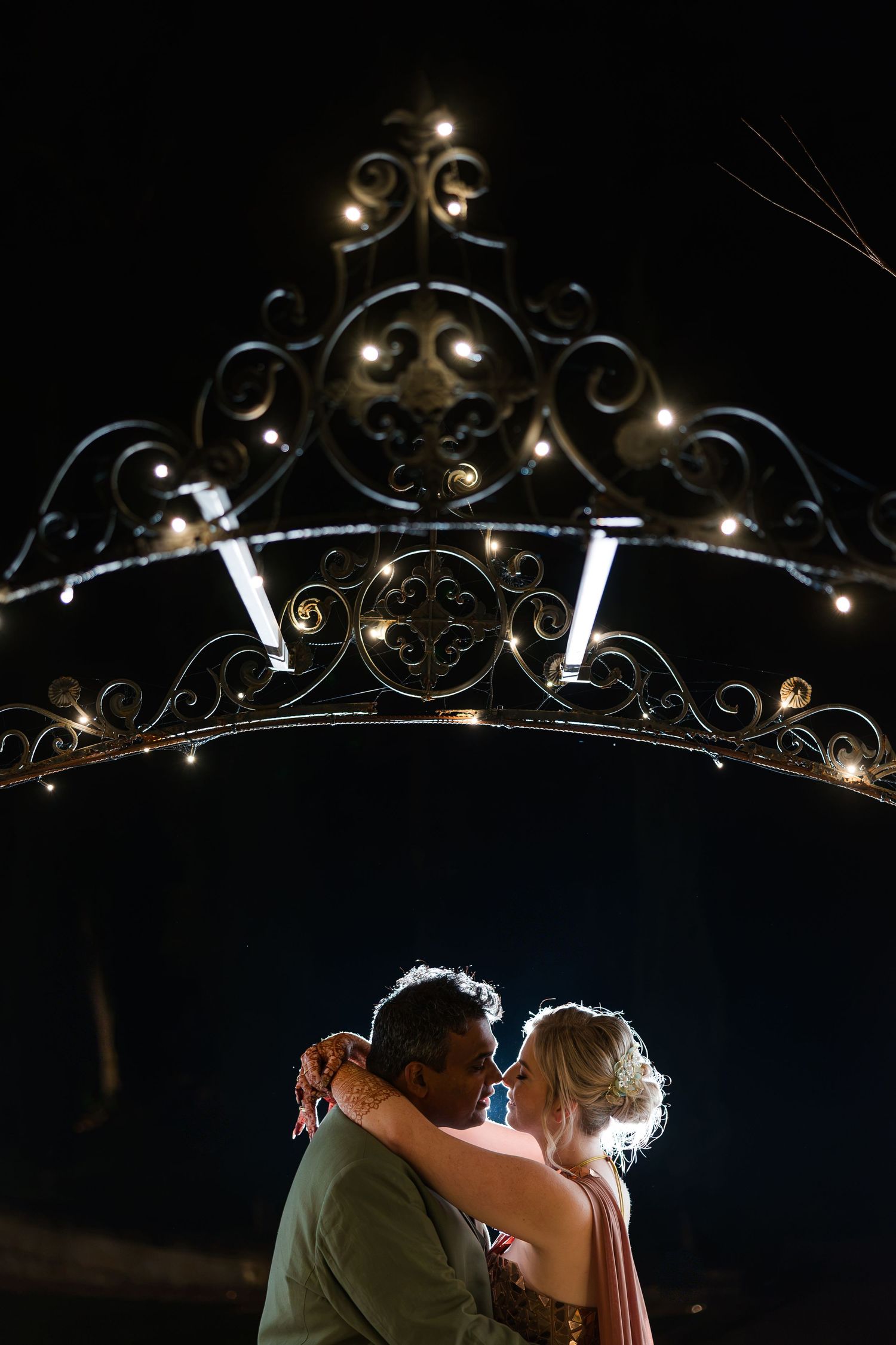 Couple embracing romantically at night beneath ornate iron arch decorated with fairy lights.