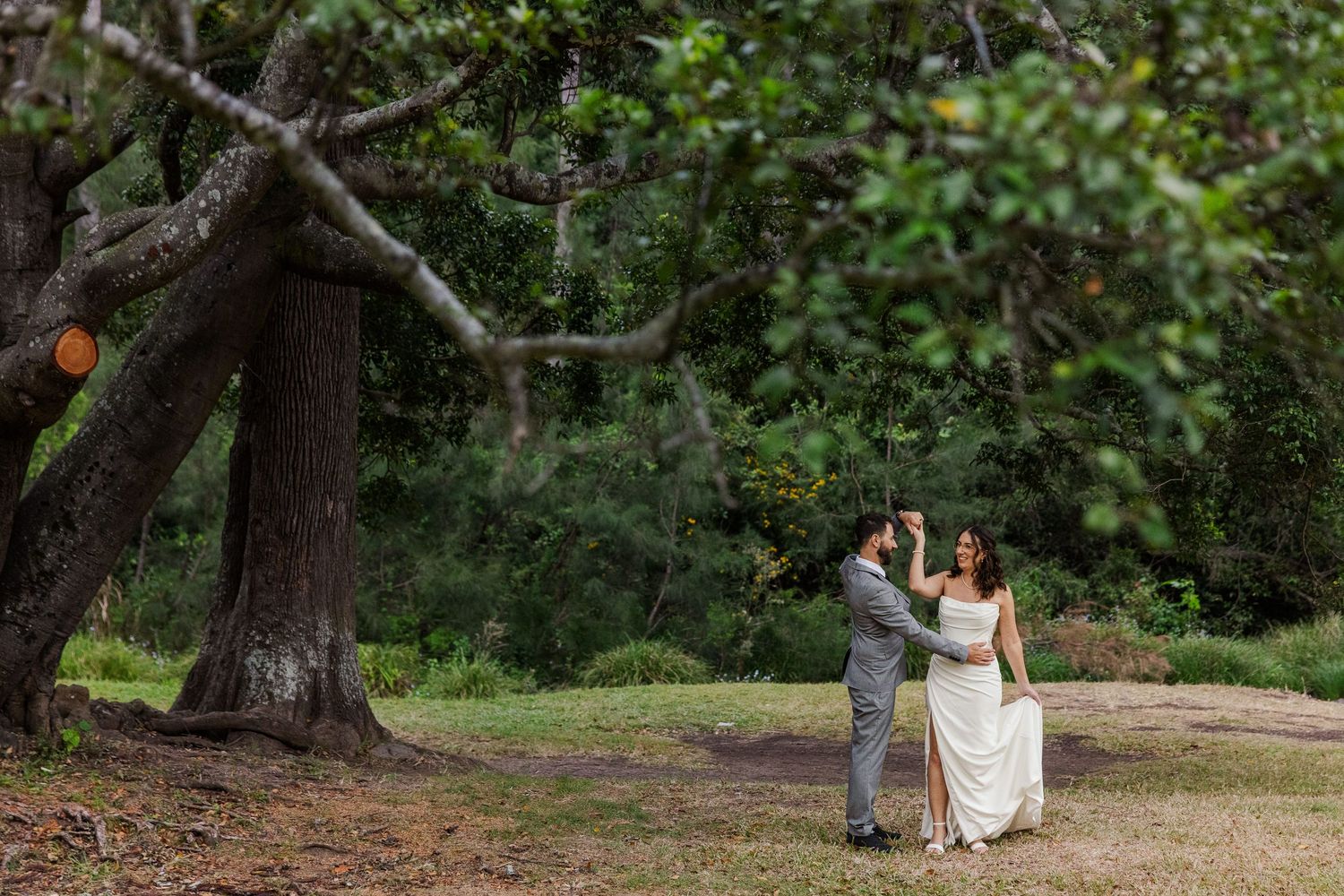A bride and groom share a romantic first dance outdoors surrounded by lush green trees in a forest setting.