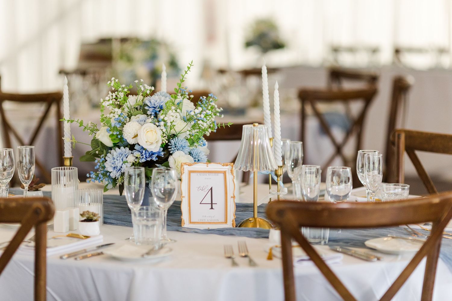 Elegant wedding reception table with blue hydrangea centerpiece, white candles, crystal glasses, and table number 4 on white linen.