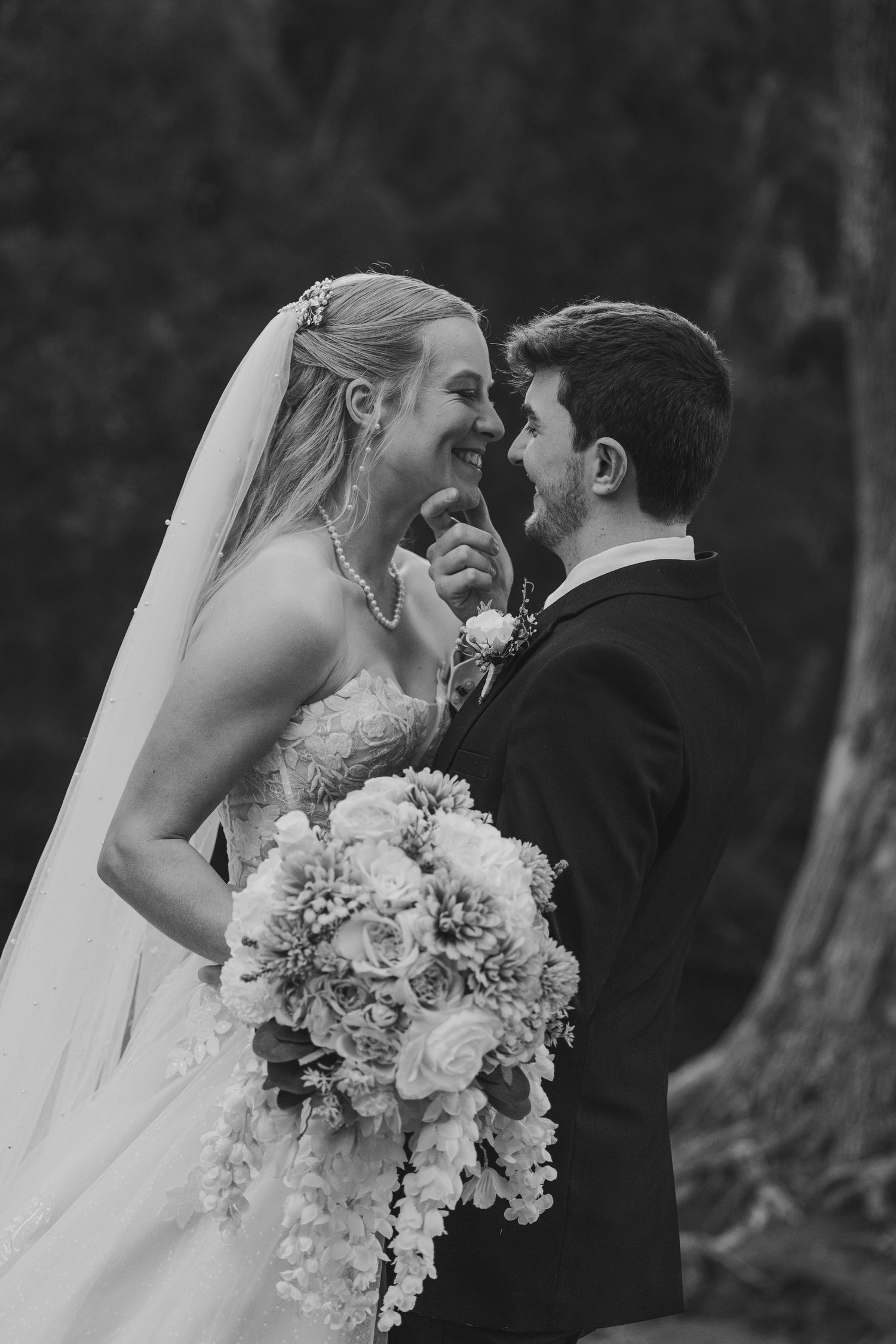 Black and white photo of a laughing bride holding a floral bouquet and groom in a tuxedo sharing a joyful moment.
