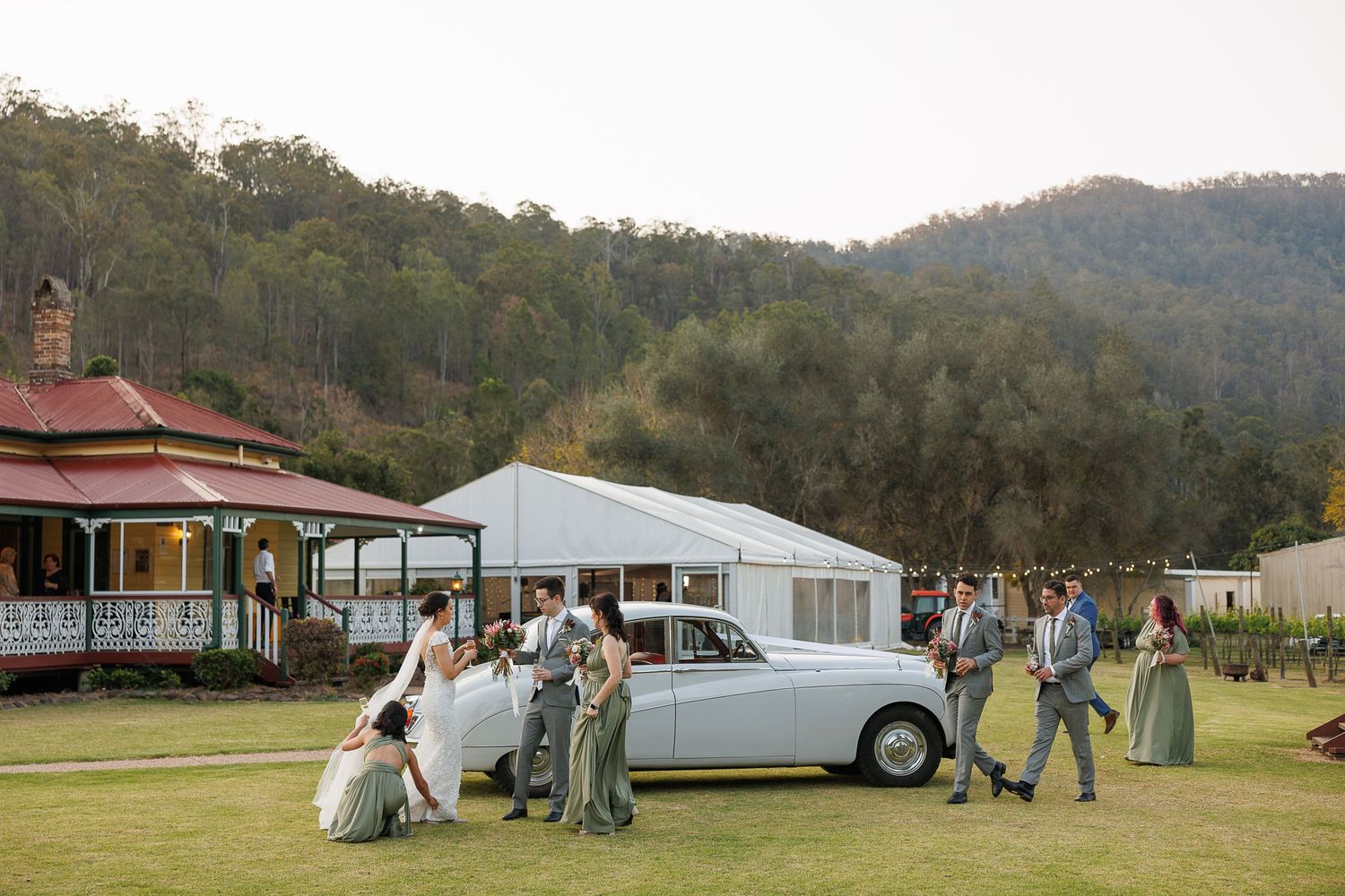 Wedding party poses beside a vintage white car on green lawn with mountain backdrop and marquee tent in Australia.