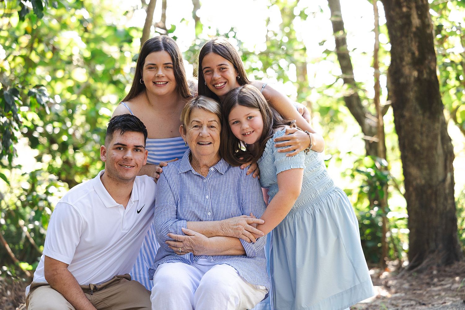 A loving family embraces while seated together outdoors in a sunny wooded setting wearing blue and white clothing.
