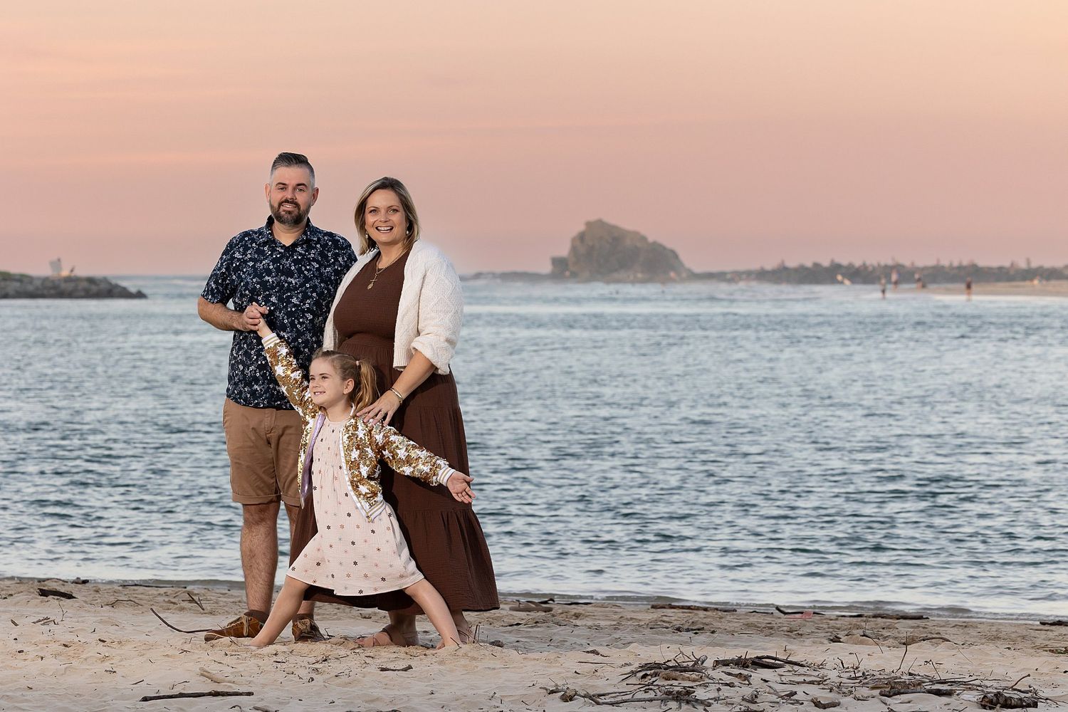 A family stands together at sunset on a beach with a rocky island visible in the distance over calm waters.