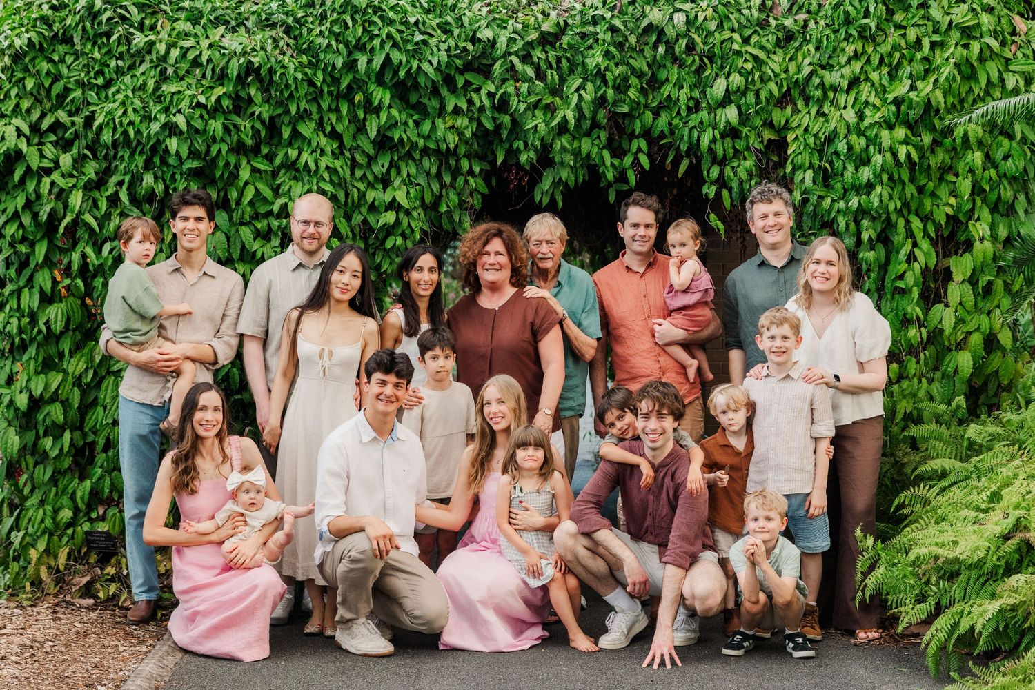 Large group wearing casual attire poses together outdoors against lush green foliage background.