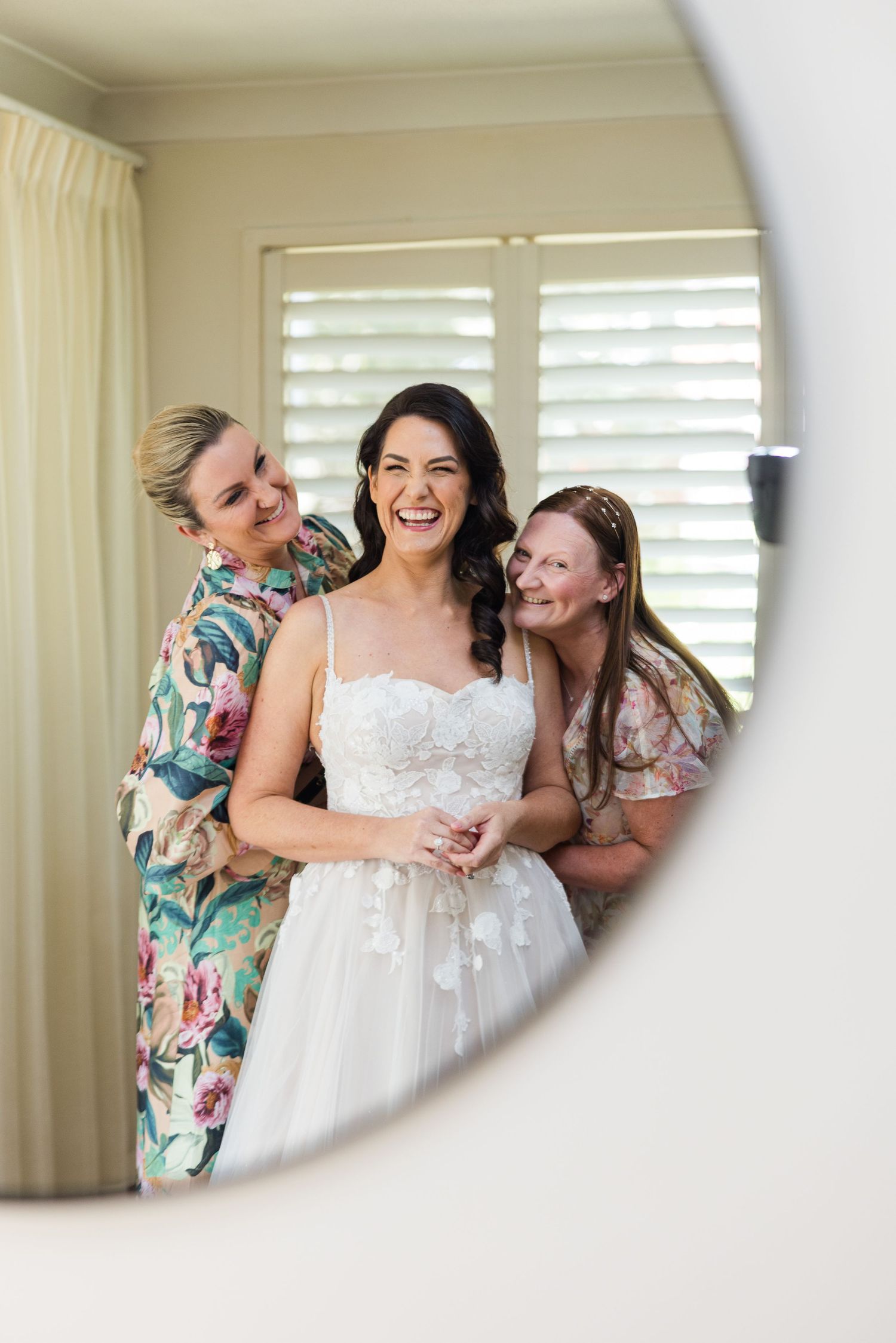Three people share a joyful moment in a sunlit room with white shutters and a beautiful white lace wedding dress.