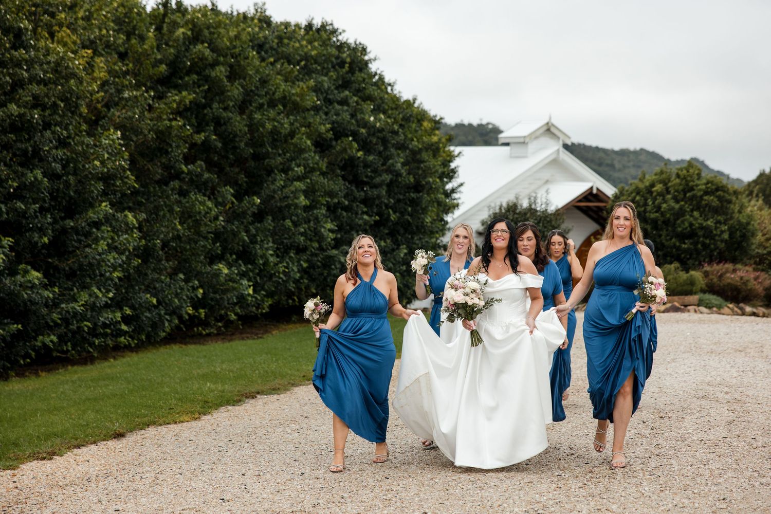 Bride in white gown walks with bridesmaids in blue dresses on gravel path near white chapel and lush greenery.