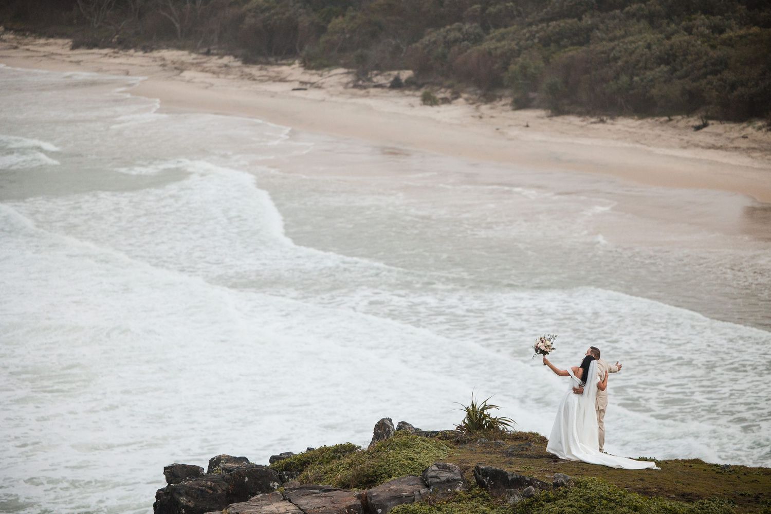 Two brides celebrate on a rocky cliff overlooking a stunning beach with rolling waves during their wedding day.