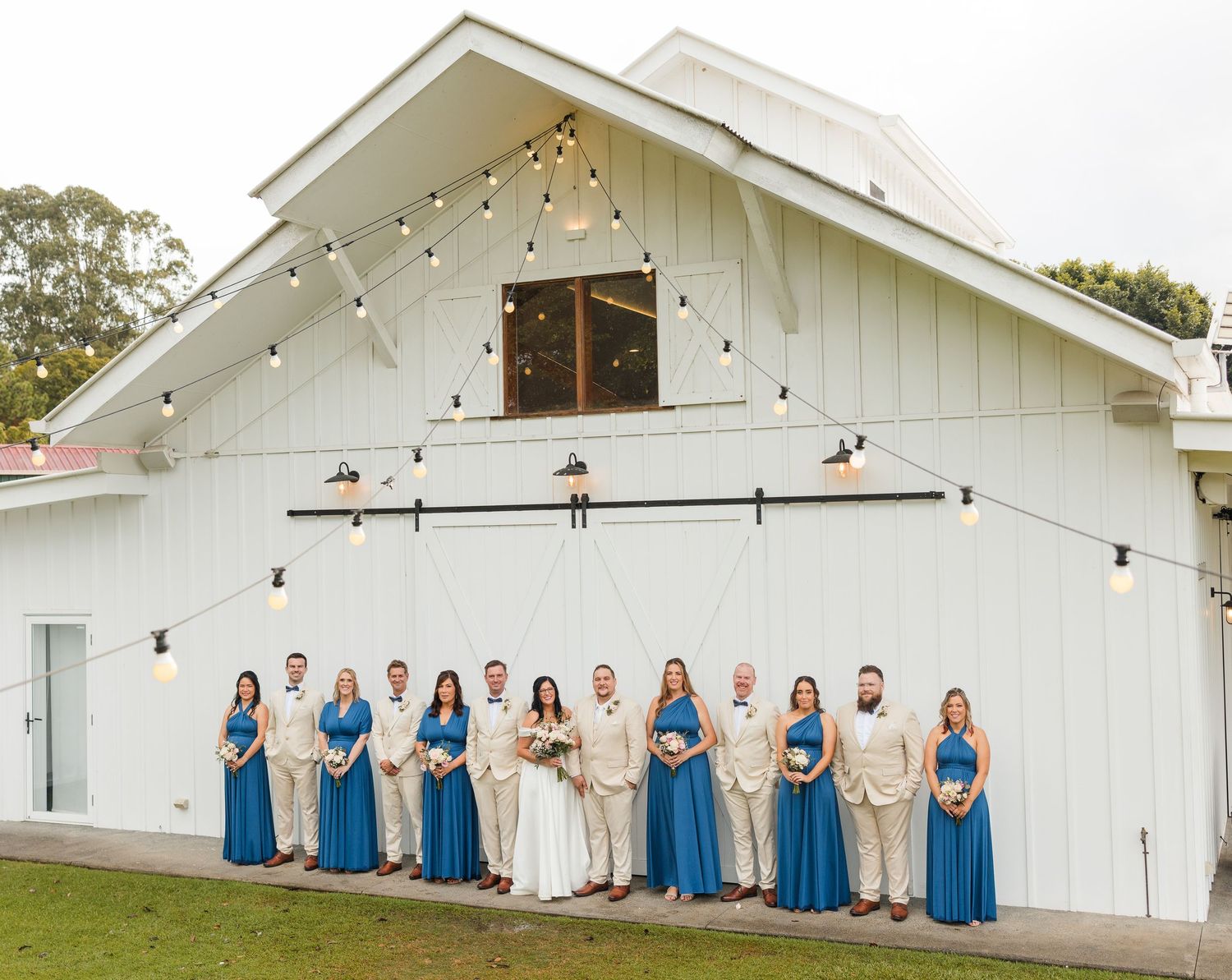 Wedding party poses in blue and tan attire before a white barn venue with string lights.