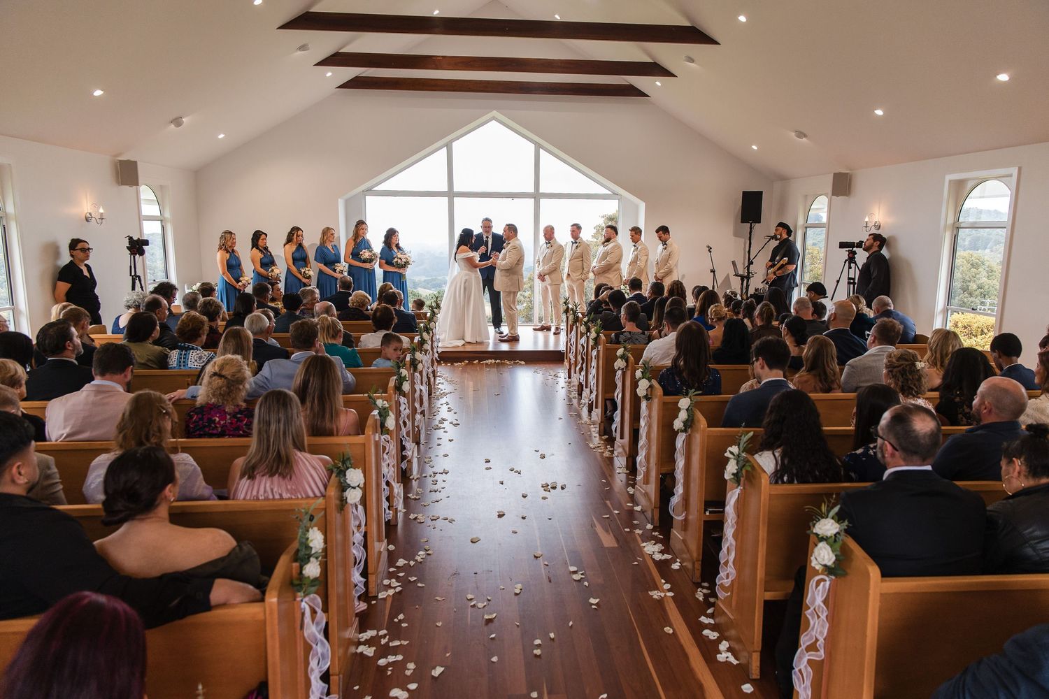 Wedding ceremony in a bright chapel with bridesmaids in blue, guests in pews, and rose petals lining the aisle.