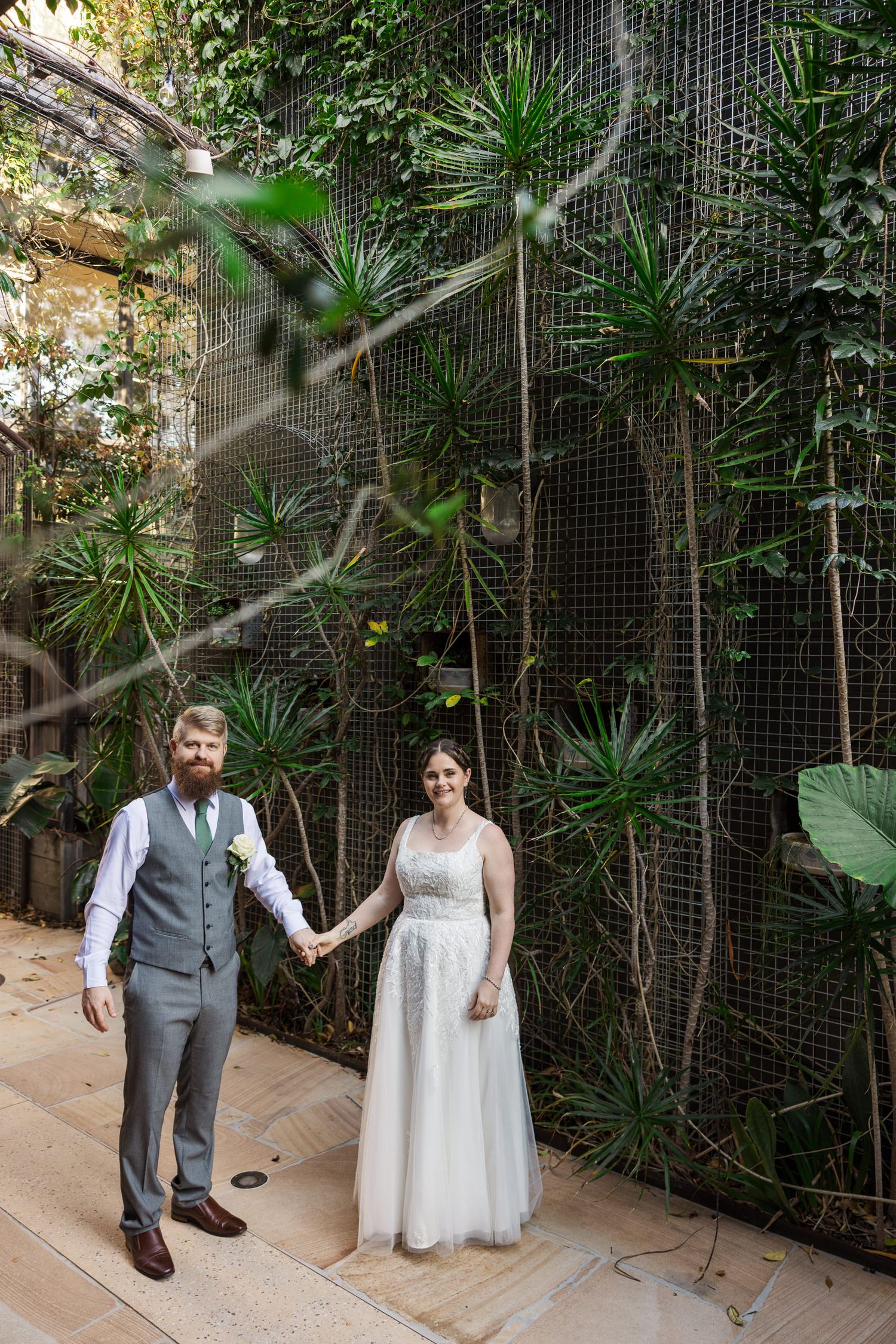 Bride and groom holding hands in front of a lush green plant wall in a botanical garden wedding setting.