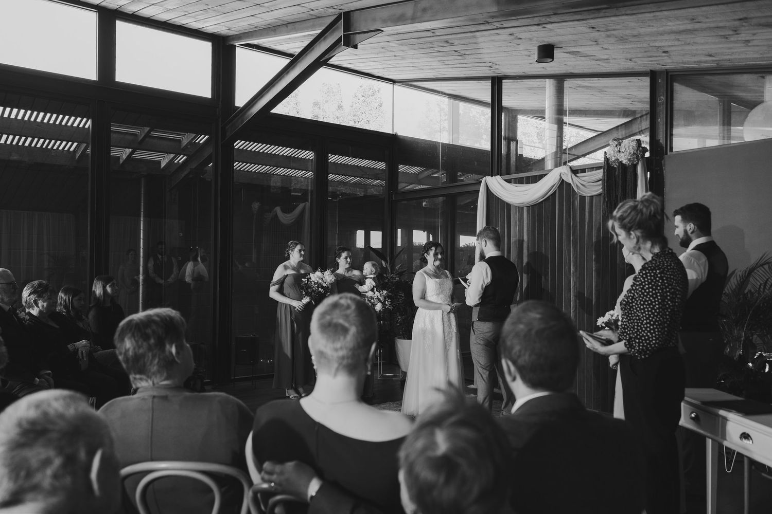 Black and white photo of an intimate indoor wedding ceremony with guests seated and bridal party at the altar.
