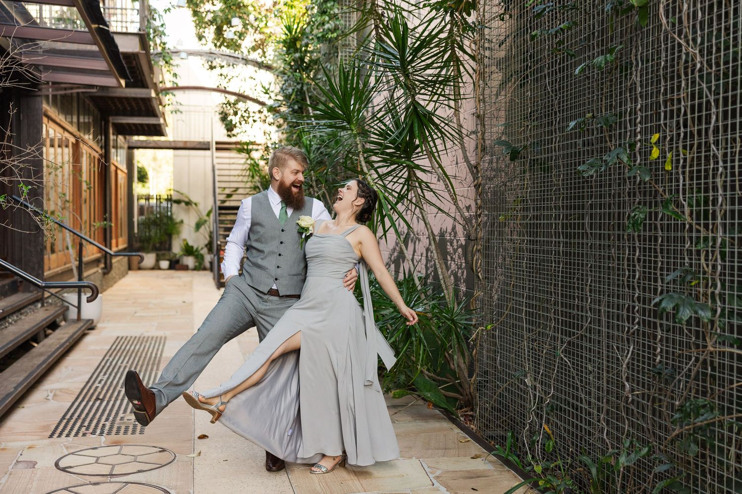 A joyful couple dances outdoors in gray wedding attire, surrounded by lush greenery and rustic walls.