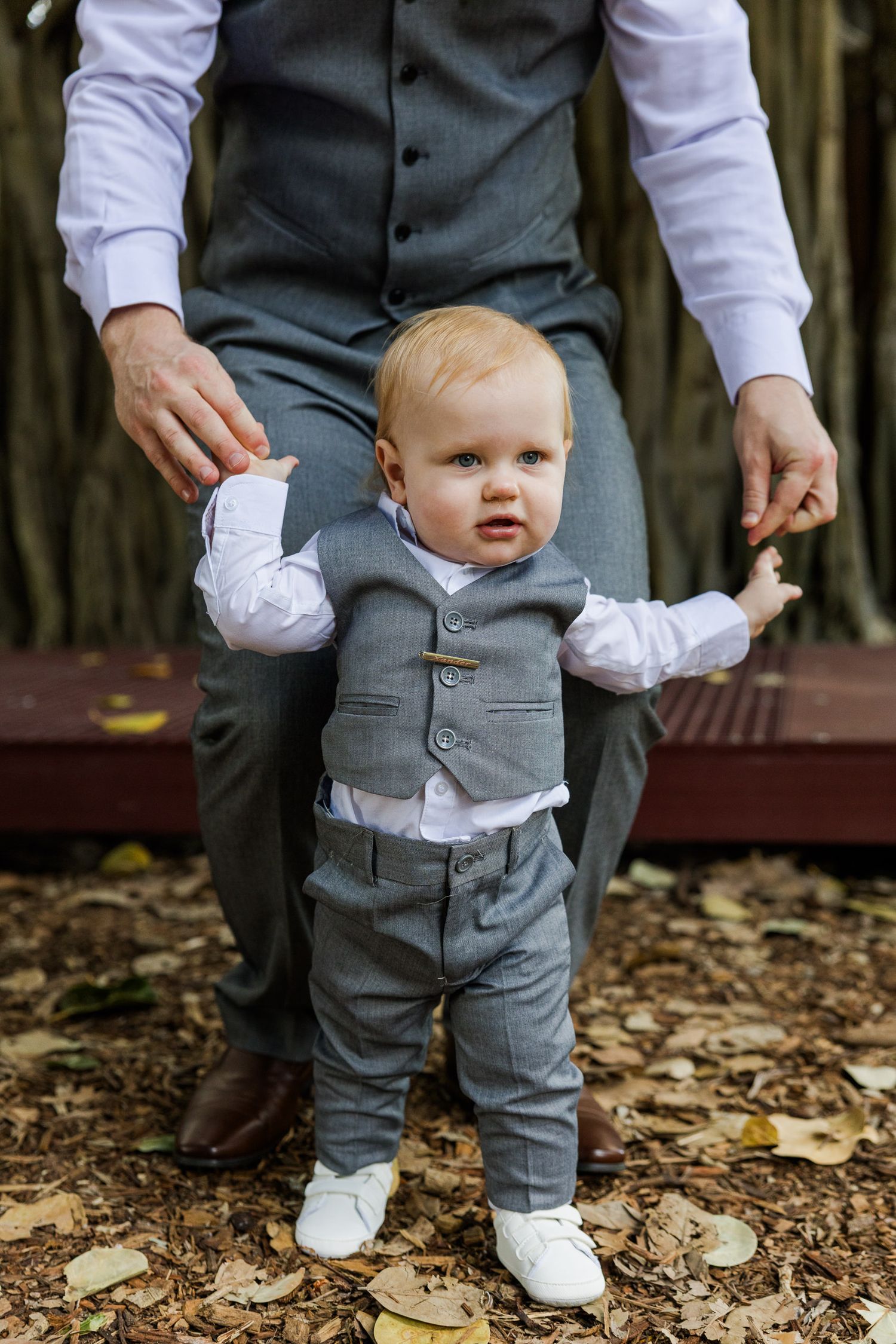 A baby boy in a matching gray vest suit stands with an adult holding his hands on a leaf-covered ground.