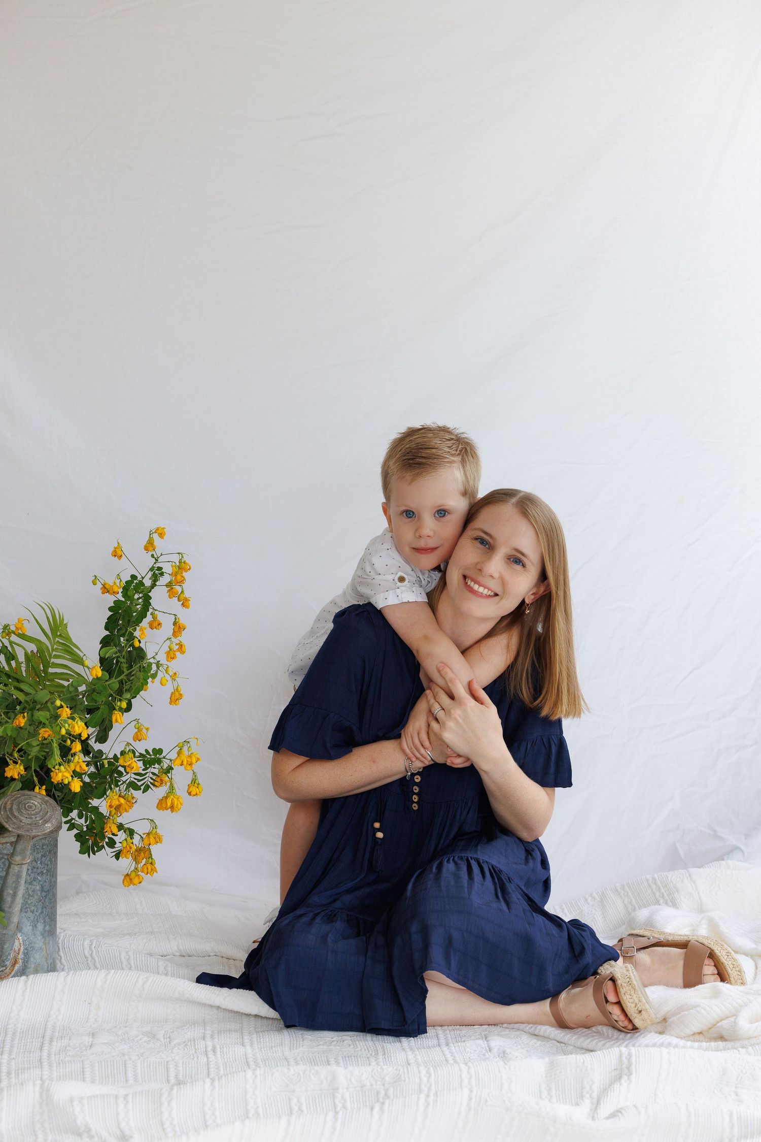 A smiling woman in a navy dress sits on a white bed while a young blonde boy hugs her from behind near yellow flowers.