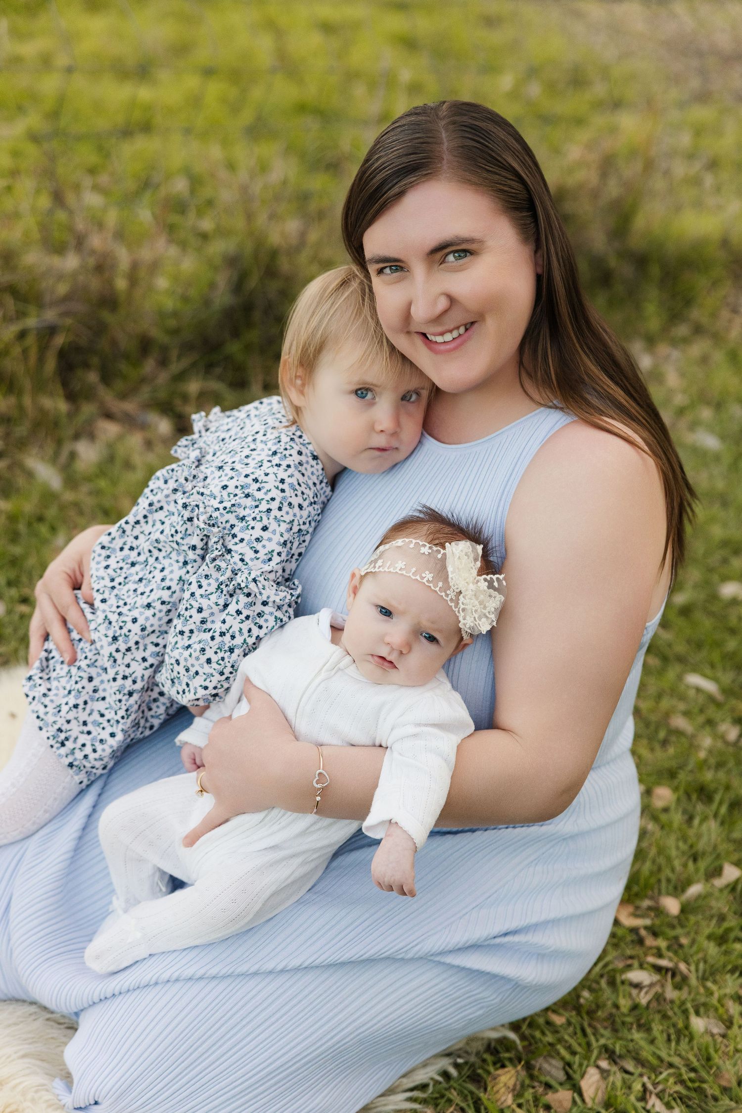A smiling mother in a blue dress sits on grass outdoors, lovingly holding her two young daughters.