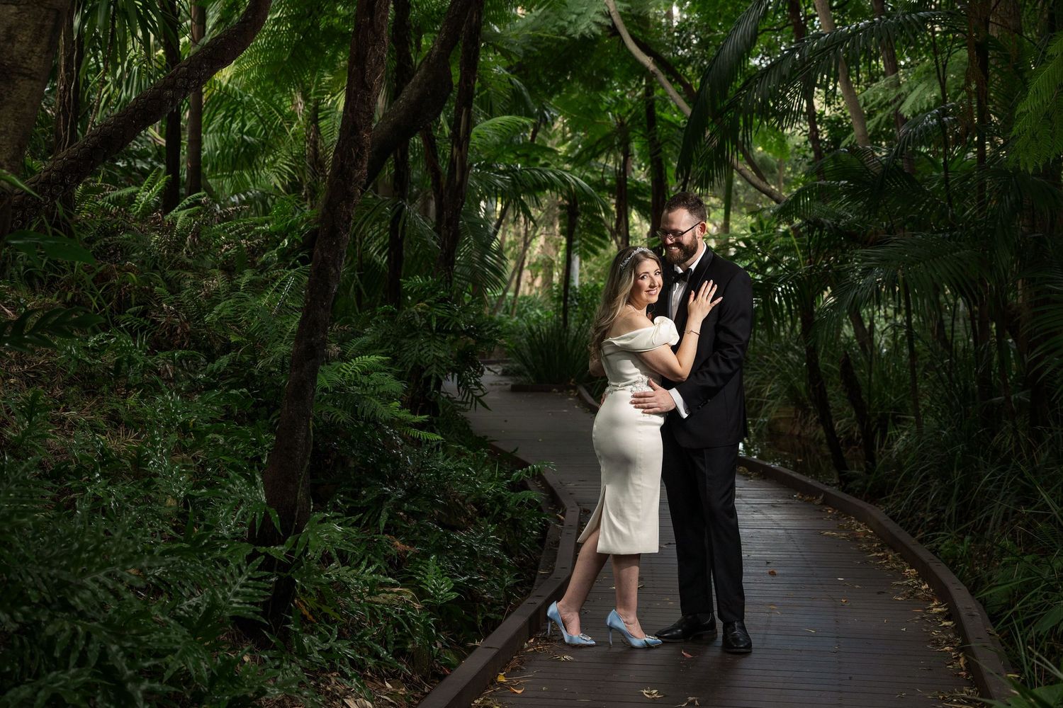 A couple embraces on a winding garden path surrounded by lush green foliage and trees.