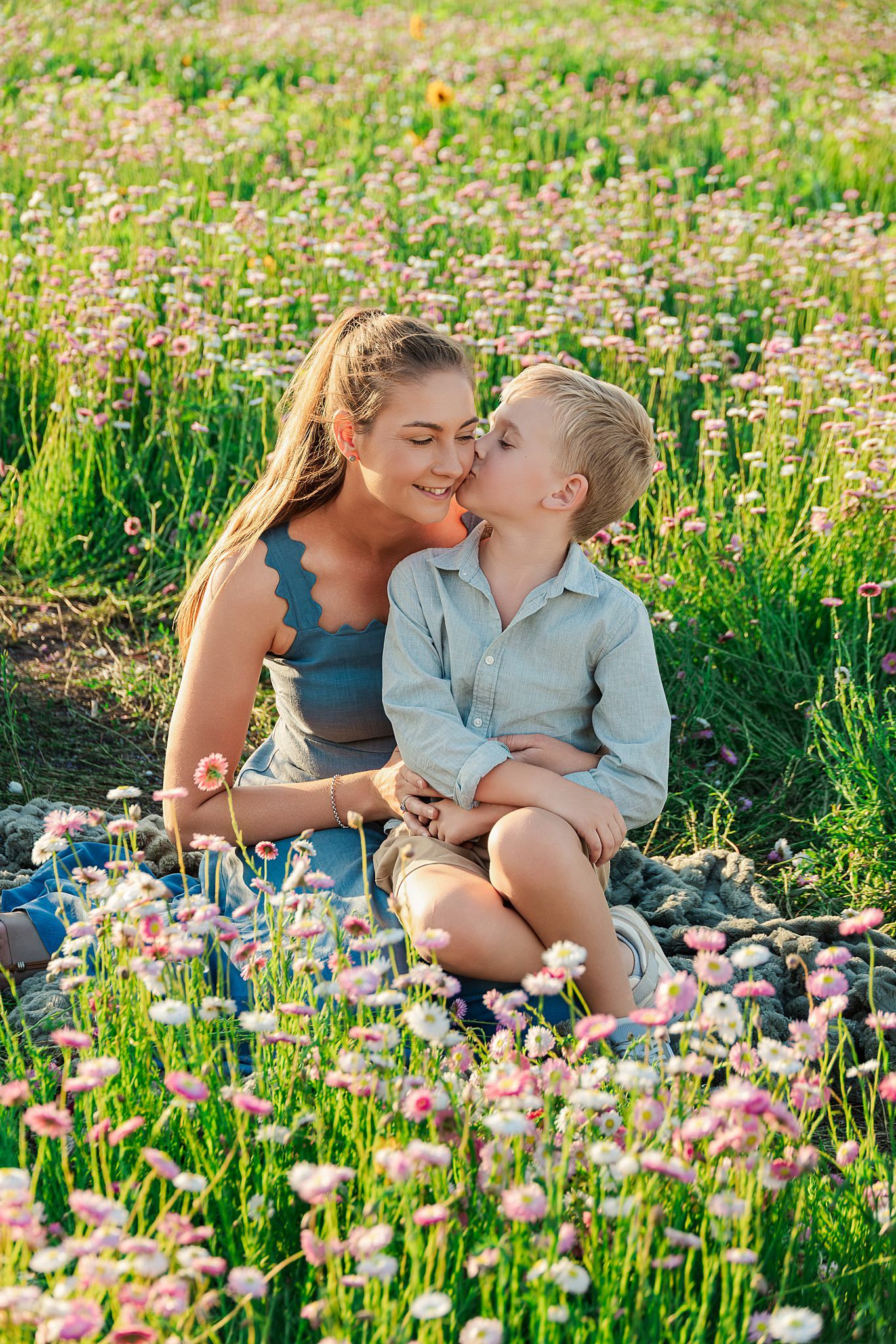 A smiling young woman holds a little boy on her lap as he kisses her cheek in a field of pink wildflowers.