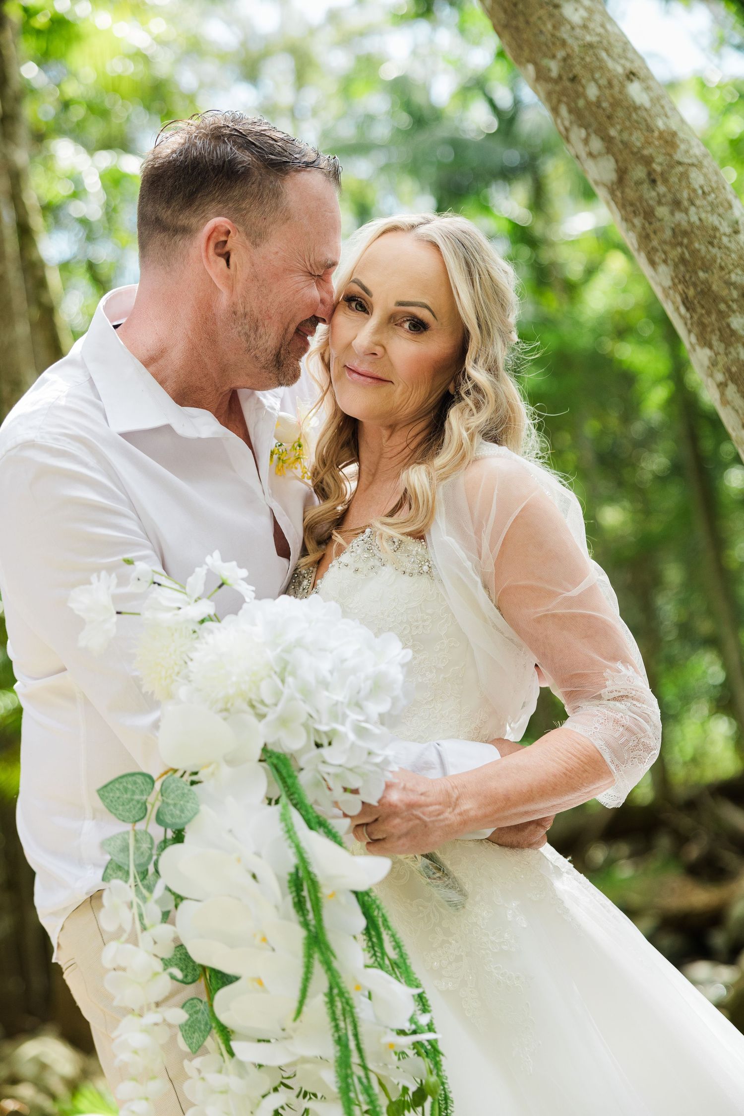 Bride and groom embrace outdoors holding white floral bouquet, smiling together in lush green garden setting.