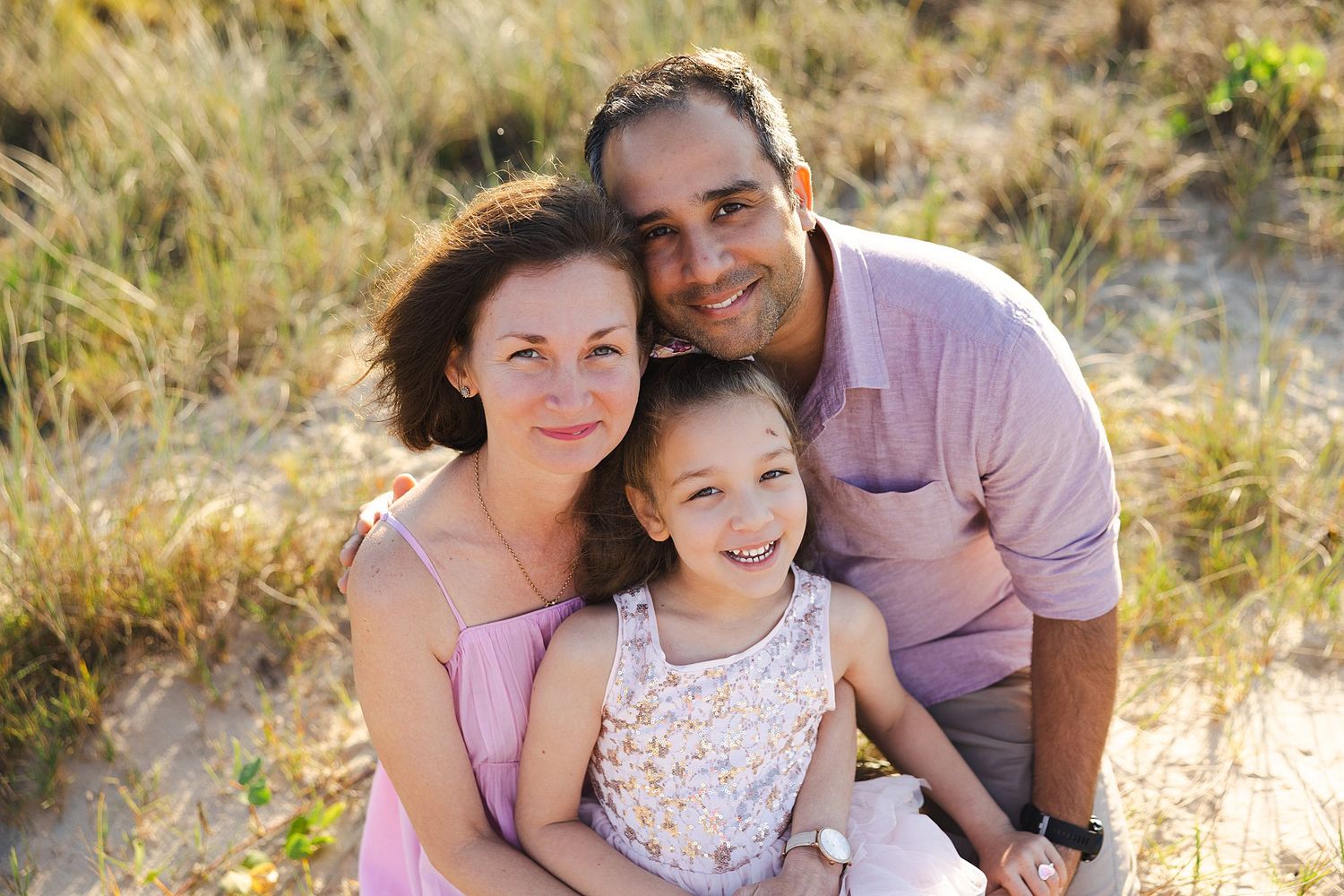 A smiling family of three poses together outdoors amid golden grasses on a sunny day.
