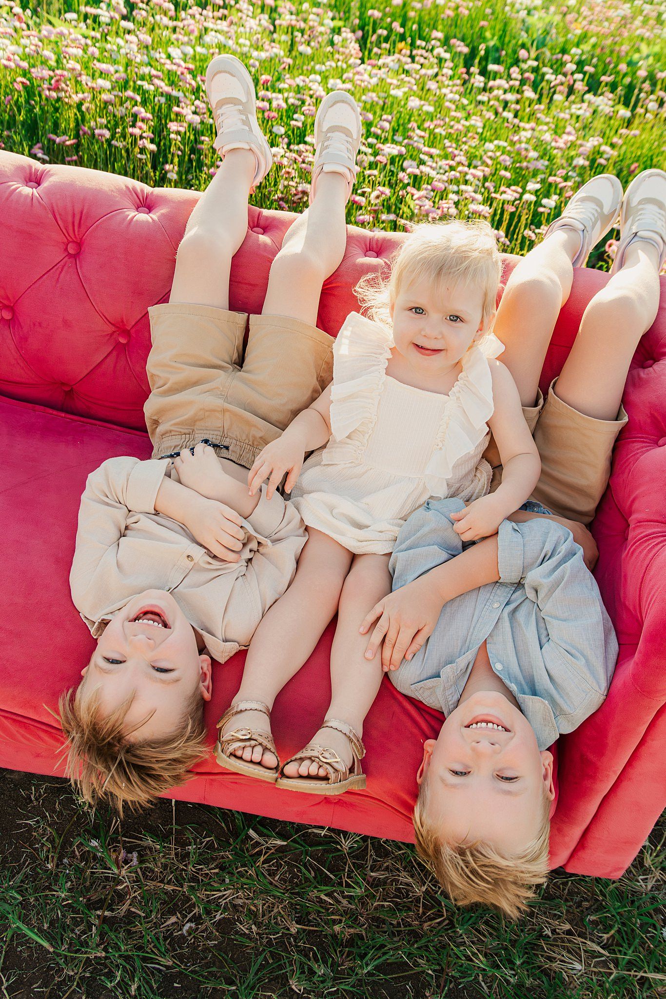 Three smiling young children in neutral outfits lie on a pink tufted sofa in a flower field, viewed from above.