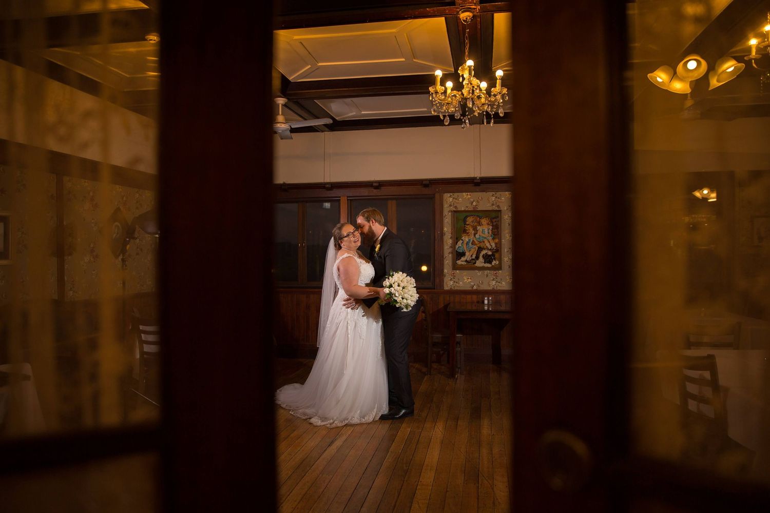 A romantic wedding couple dances in an elegant ballroom with warm lighting and chandeliers overhead.