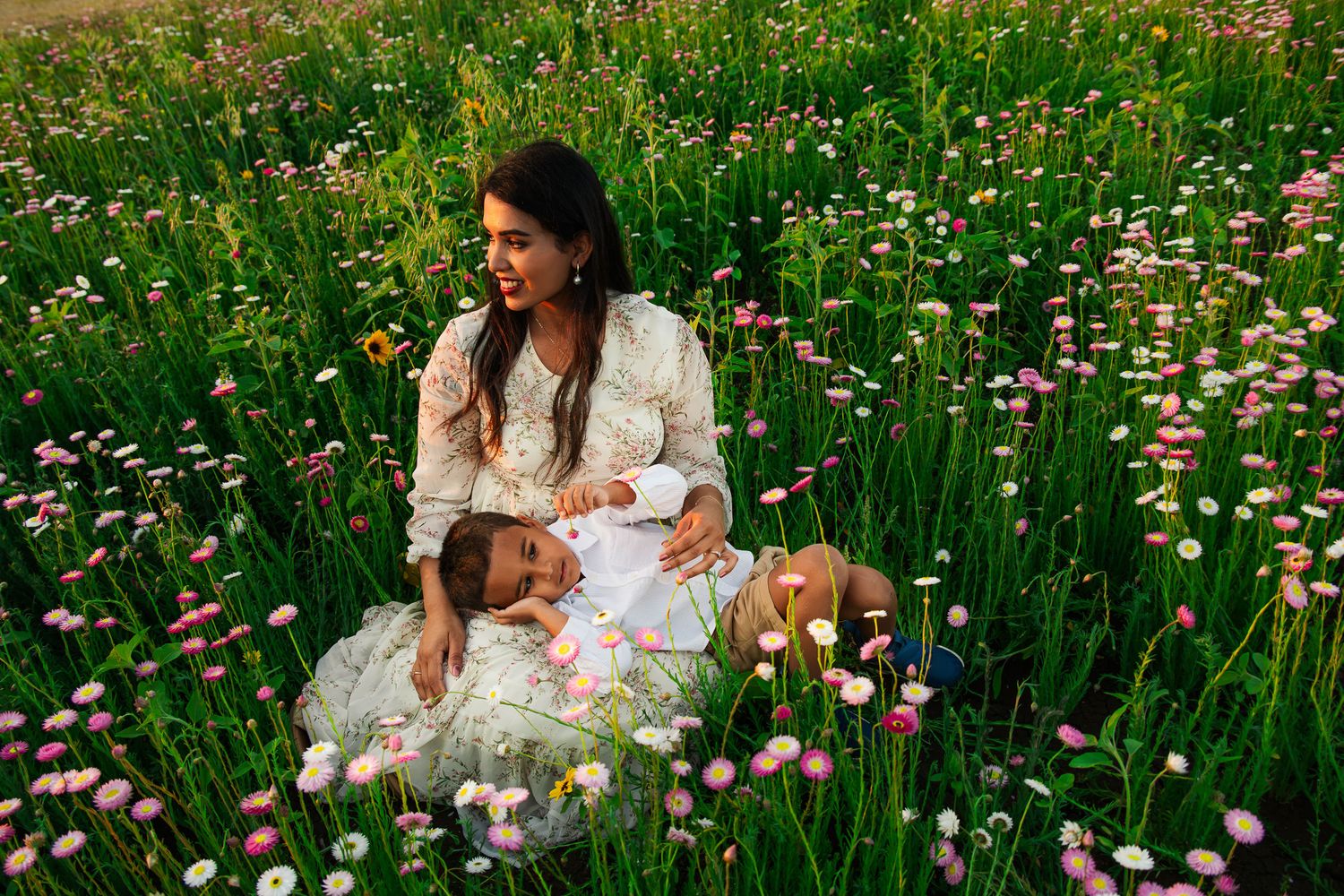 A woman in a floral dress sits with a child in a lush field of pink and white wildflowers.