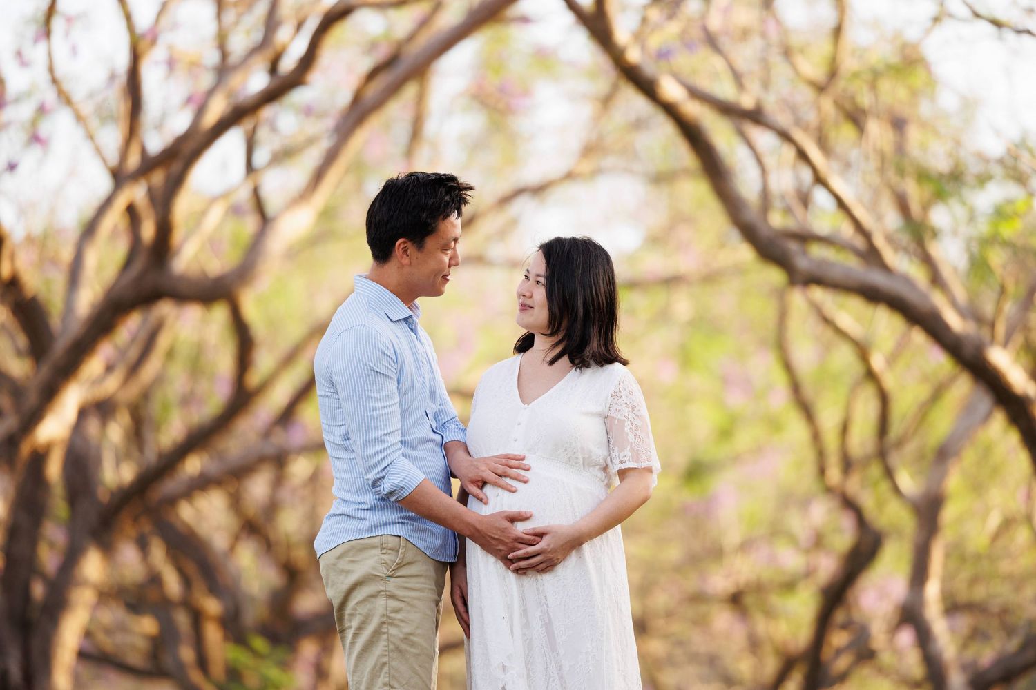 Expectant couple shares tender moment in autumn forest setting with sunlight filtering through branches.