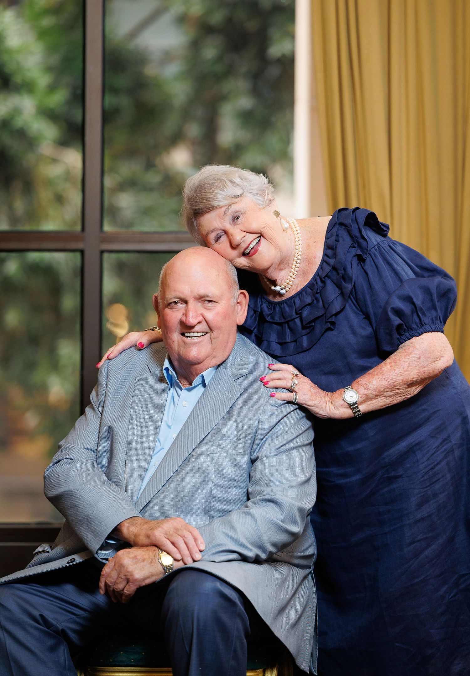 An elderly couple in formal attire sit close together by a window with curtains, sharing a warm moment.