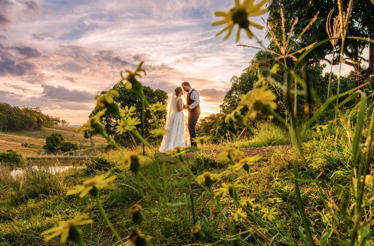 Couple stands on grassy hillside surrounded by vibrant wildflowers during a golden sunset.