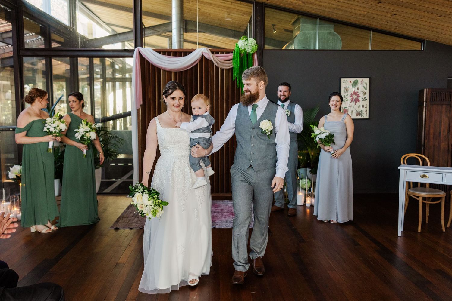 Bride and groom walk down the aisle with baby at indoor wedding ceremony, bridesmaids in green dresses in background.