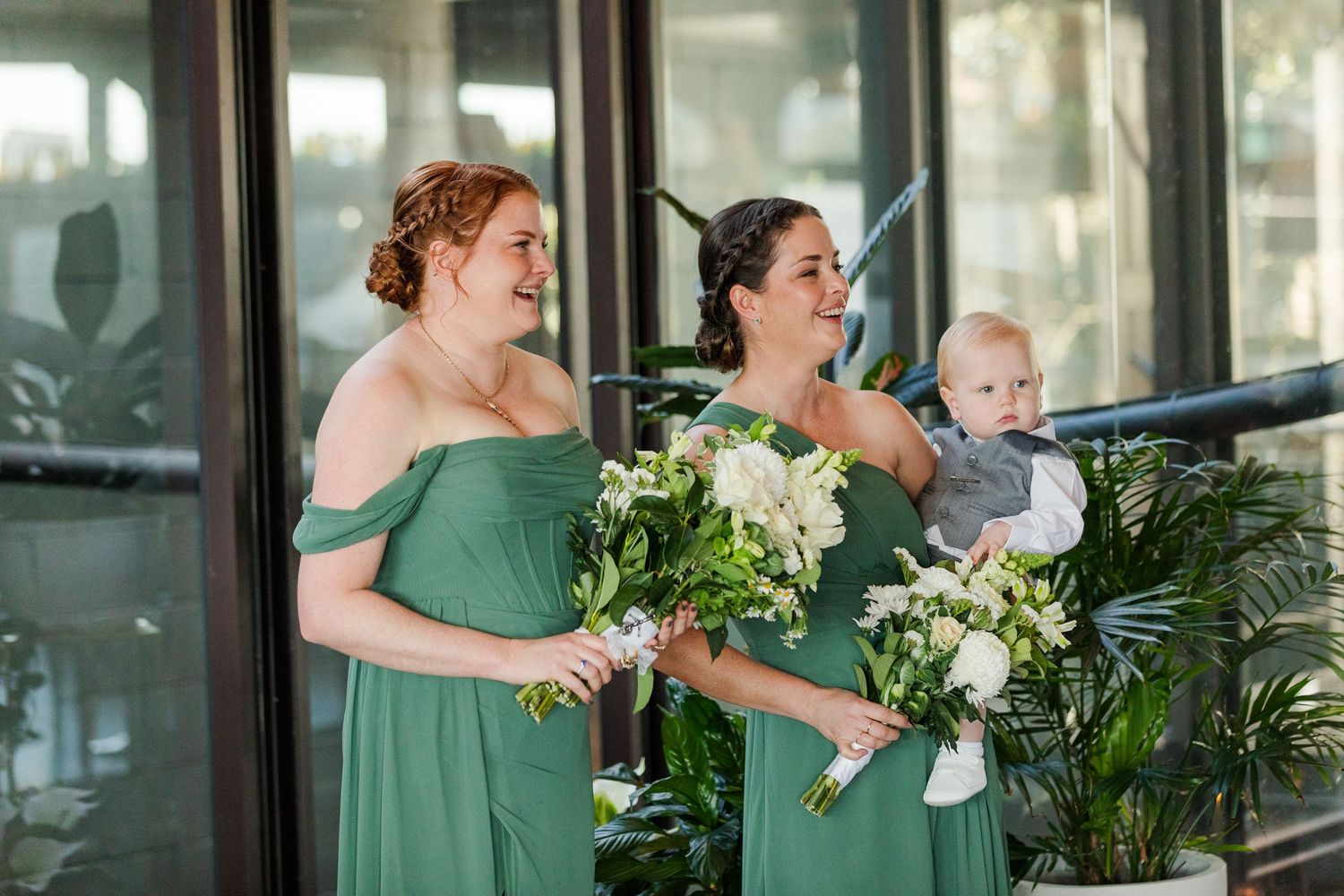 Two bridesmaids in sage green off-shoulder gowns holding white bouquets, one carrying a baby, at a wedding venue.
