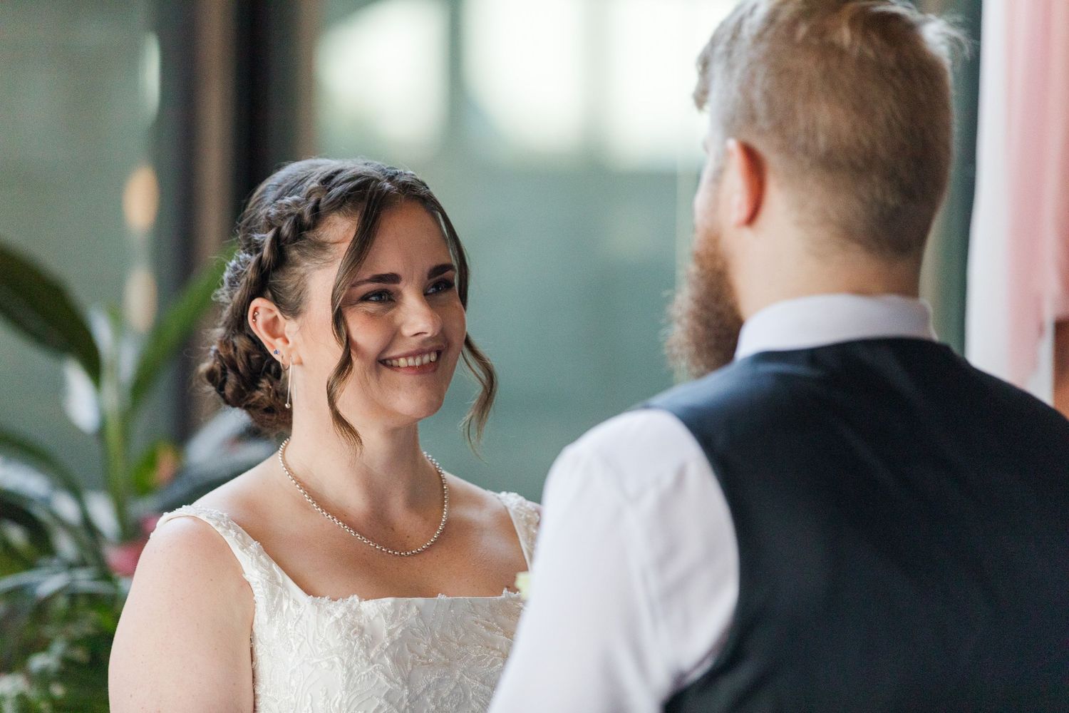 Bride smiling lovingly at groom during wedding ceremony, wearing lace dress with pearl necklace and elegant updo.