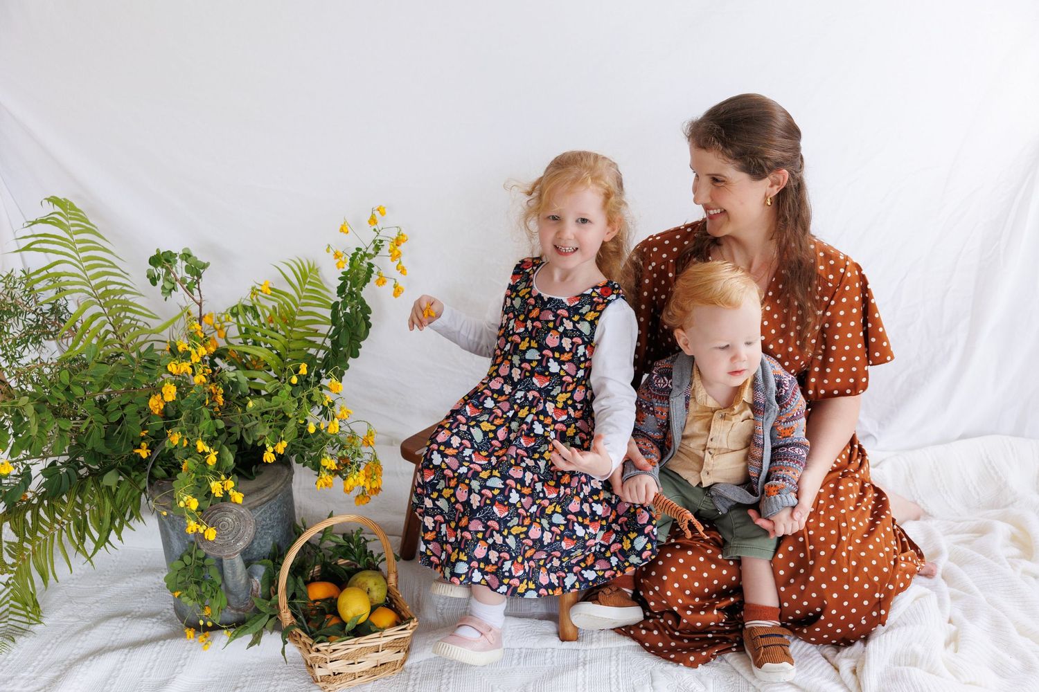 A mother sits with her two young children among yellow flowers and greenery in a bright, airy studio setting.