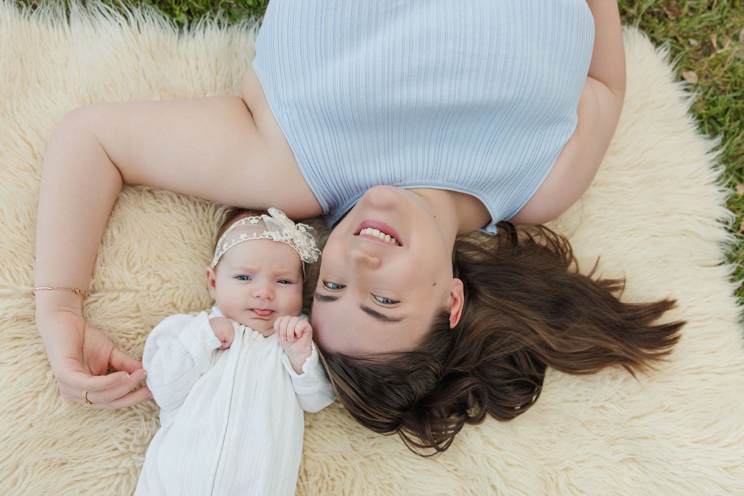 A mother and baby girl lying on a fluffy white blanket outdoors, smiling together in a sweet newborn photo session.