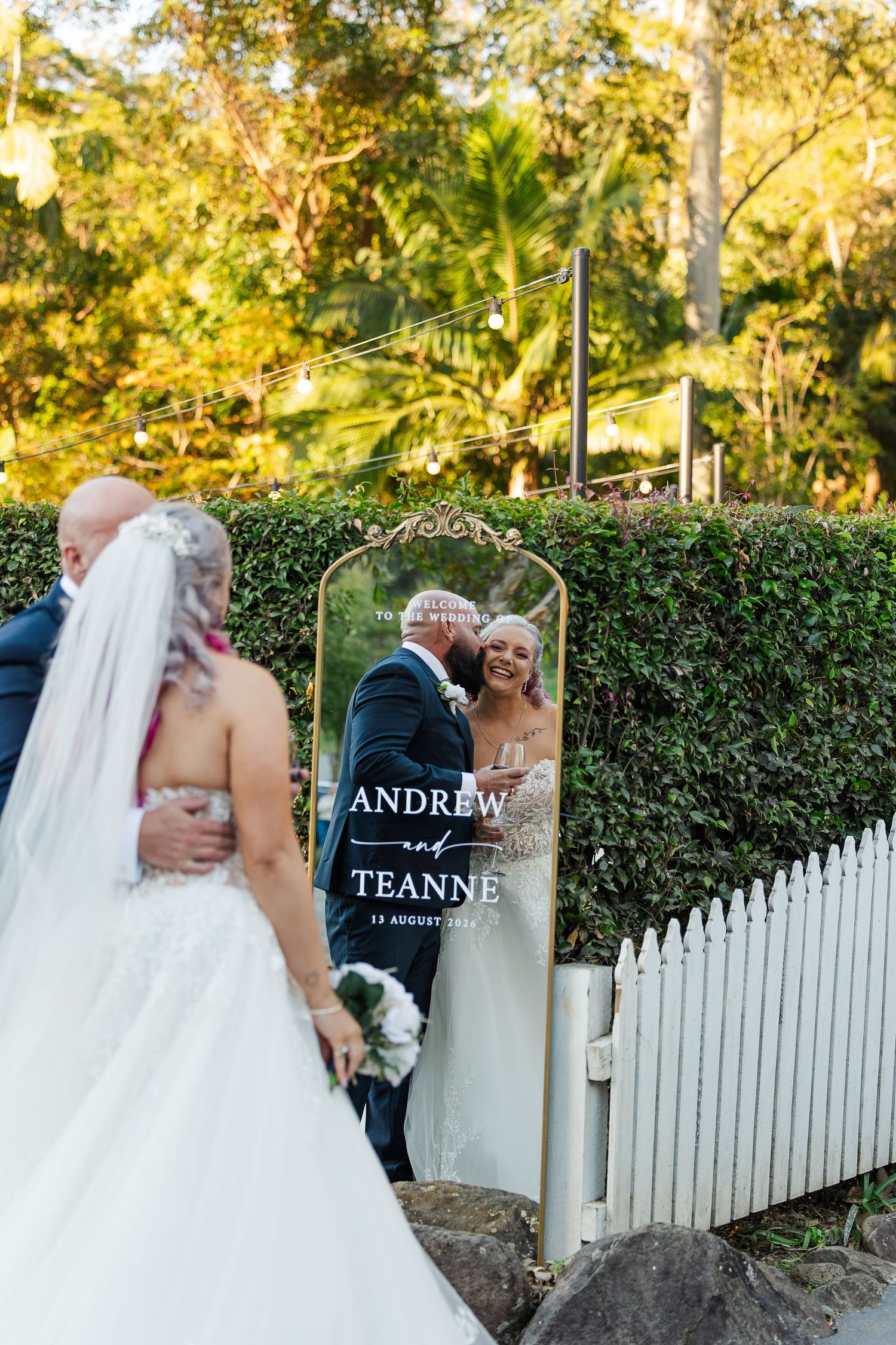 Bride and groom reflected in wedding mirror sign reading Andrew and Teanne, surrounded by lush greenery outdoors.