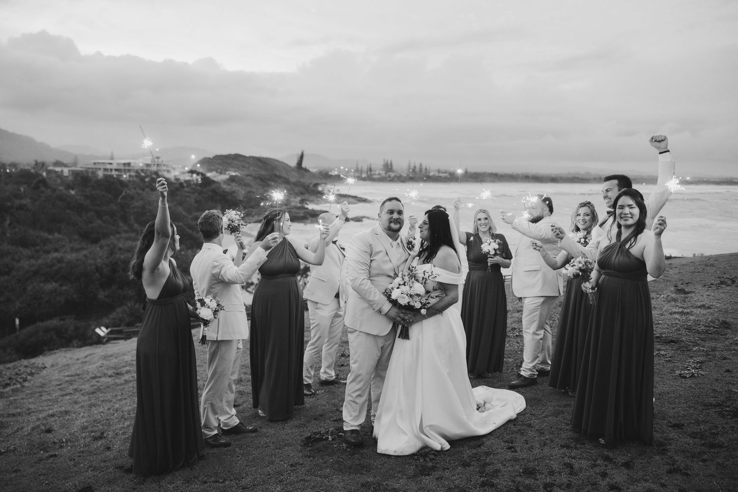 Black and white wedding photo of bride and groom surrounded by bridal party holding sparklers at an outdoor coastal ceremony.