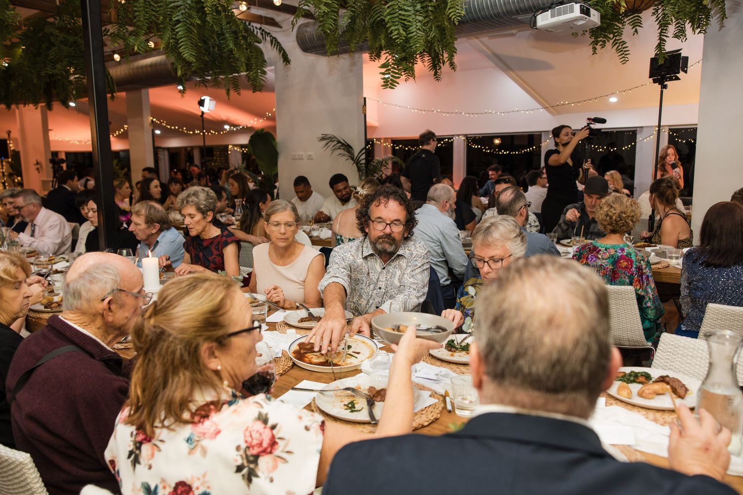 Guests dining at elegant indoor wedding reception with string lights and lush greenery decor.