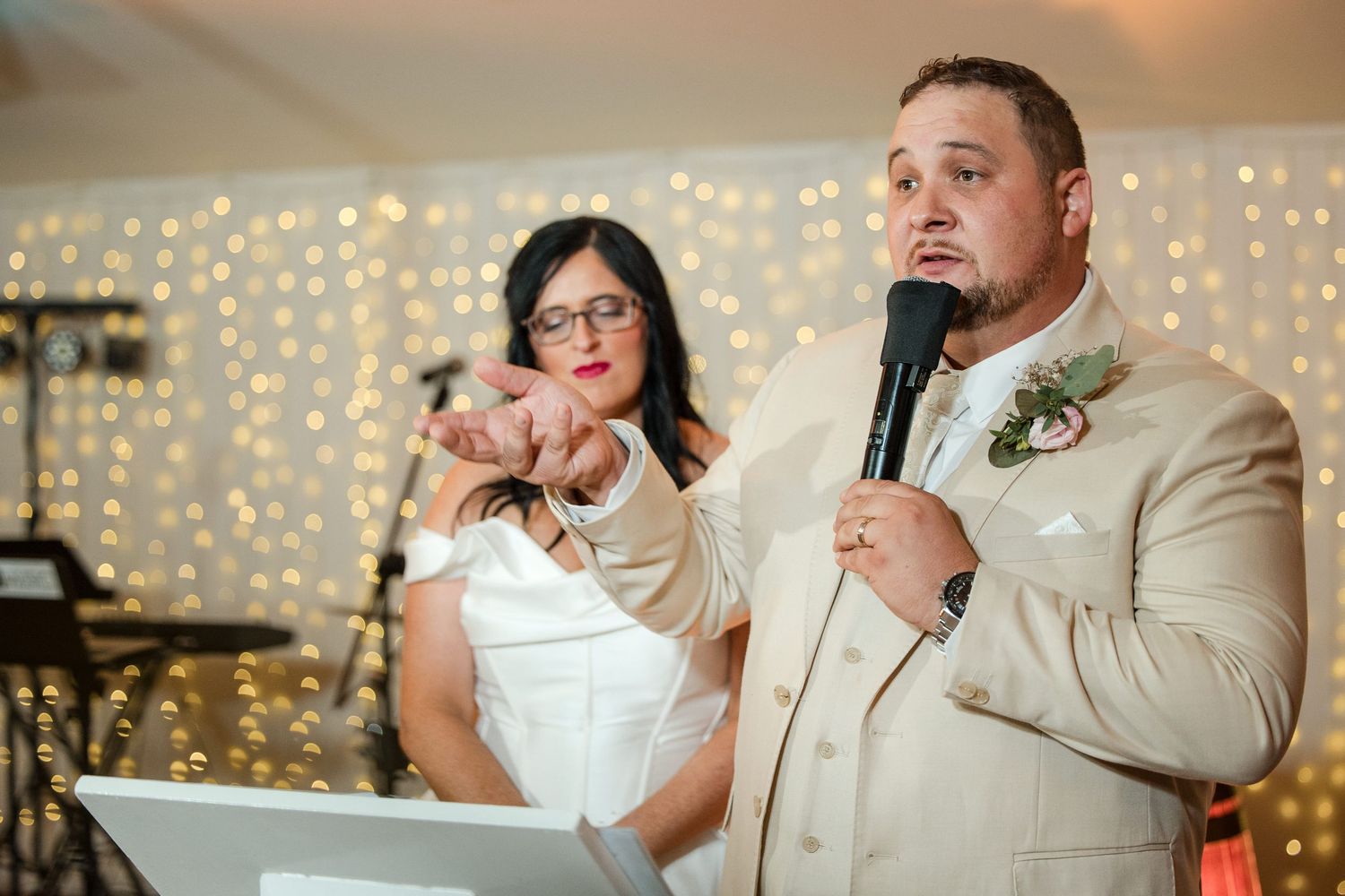 Groom in beige suit giving wedding speech with microphone, bride in background amid fairy lights.
