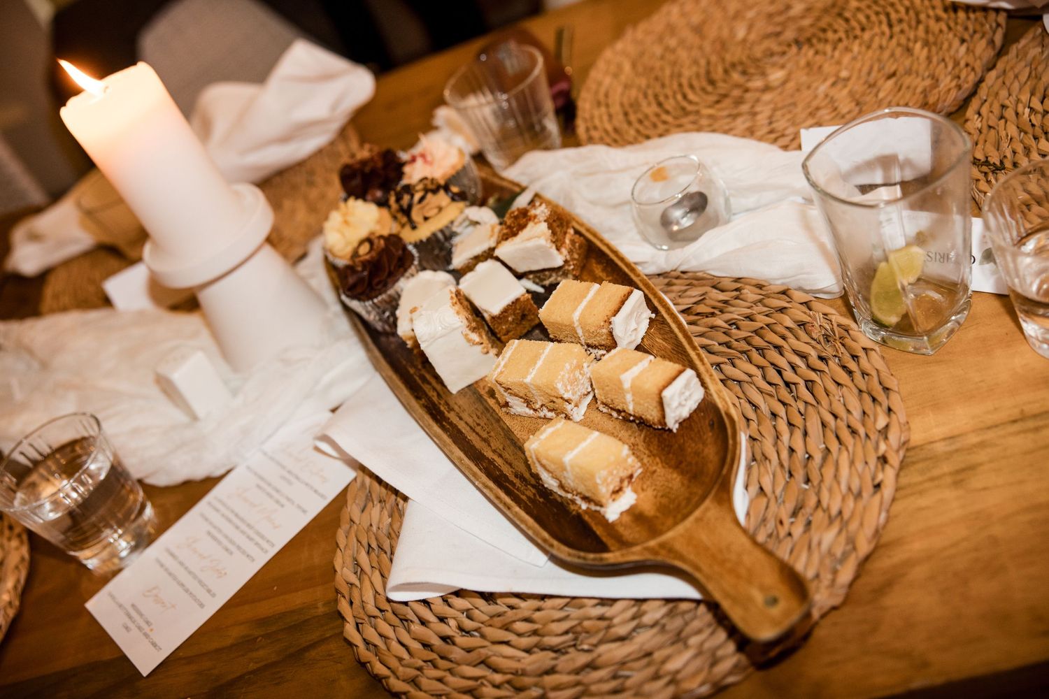 Rustic wooden serving board with assorted bread and pastries on a dining table with candles and woven placemats.