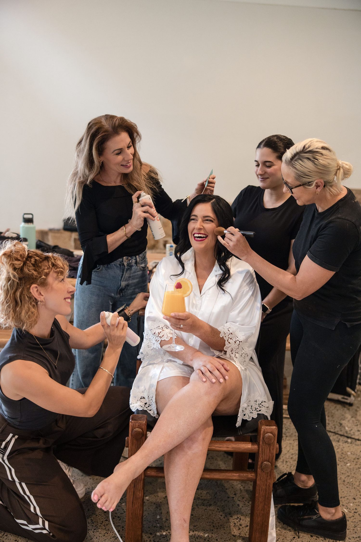 Bride in white robe laughing while four stylists apply makeup, hair, and nails during bridal getting-ready session.