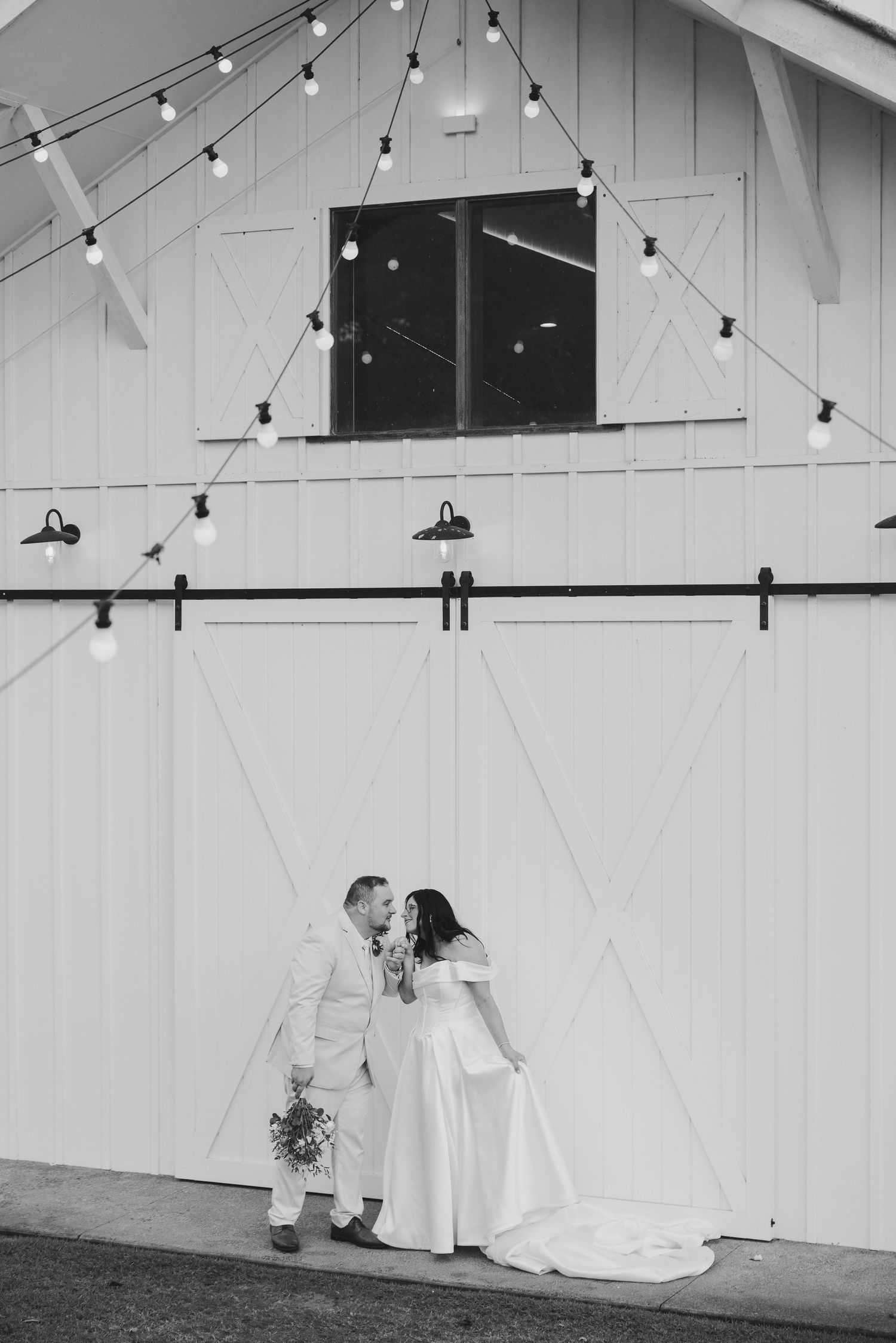 Black and white photo of a bride and groom sharing a romantic moment outside a white barn with string lights.