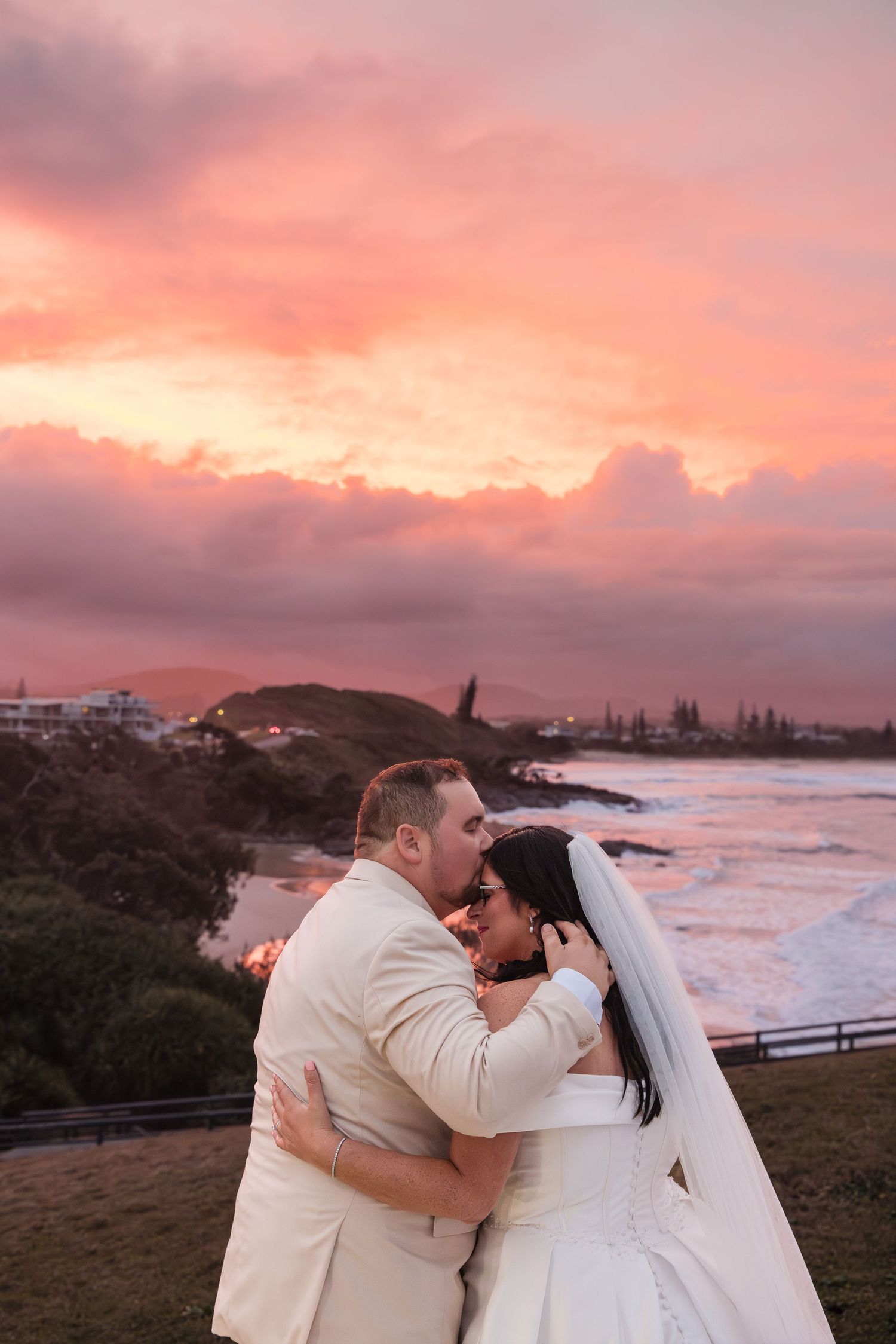 Bride and groom share romantic moment at sunset with pink cloudy sky and ocean backdrop on their wedding day.