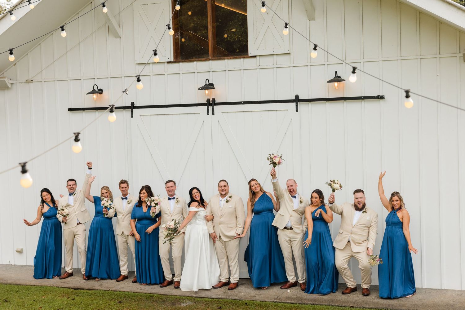 Wedding party celebrating in front of a white barn, bridesmaids in blue gowns and groomsmen in tan suits.