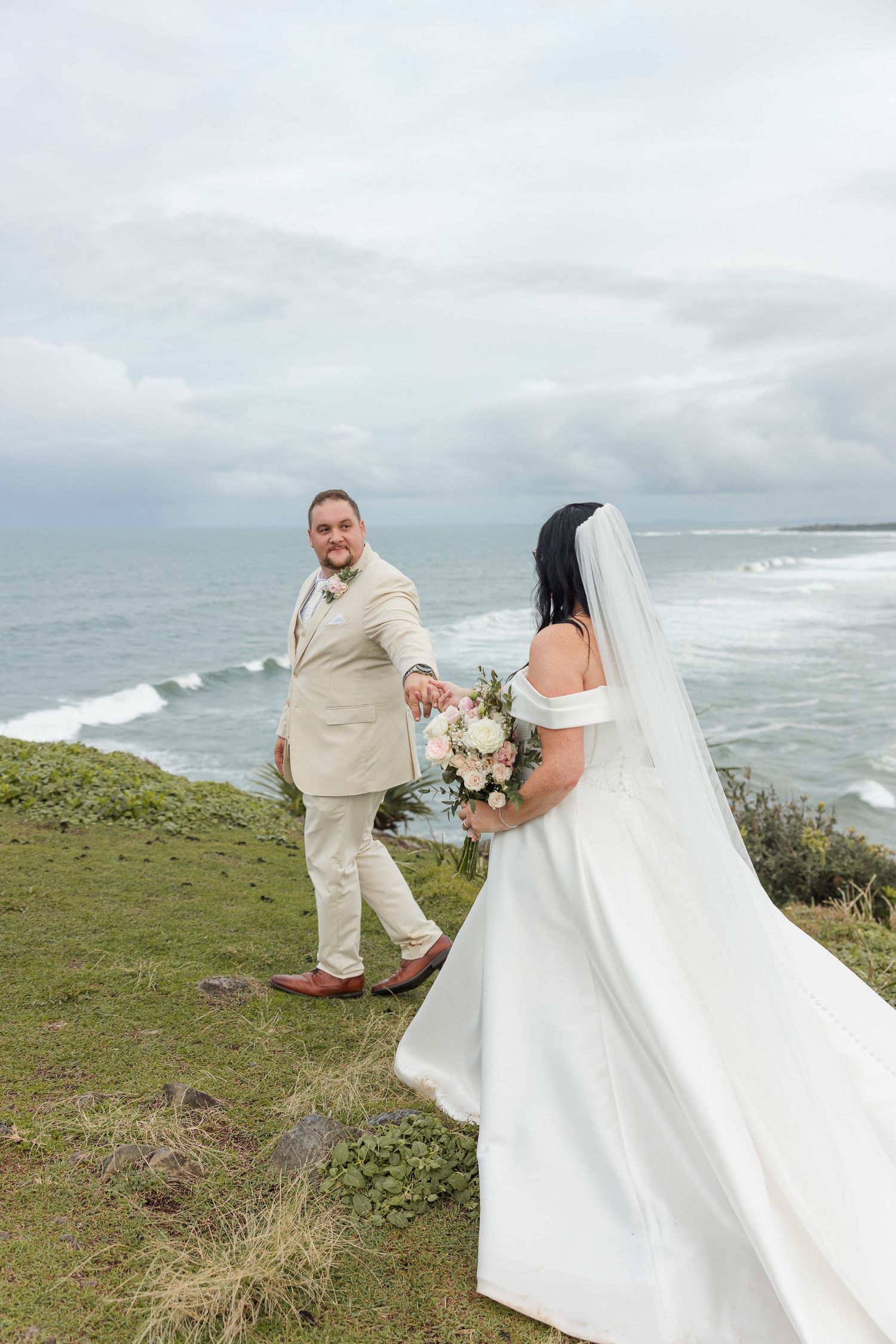 Bride and groom hold hands on a coastal cliff, bride in white gown with veil, groom in beige suit, ocean backdrop.
