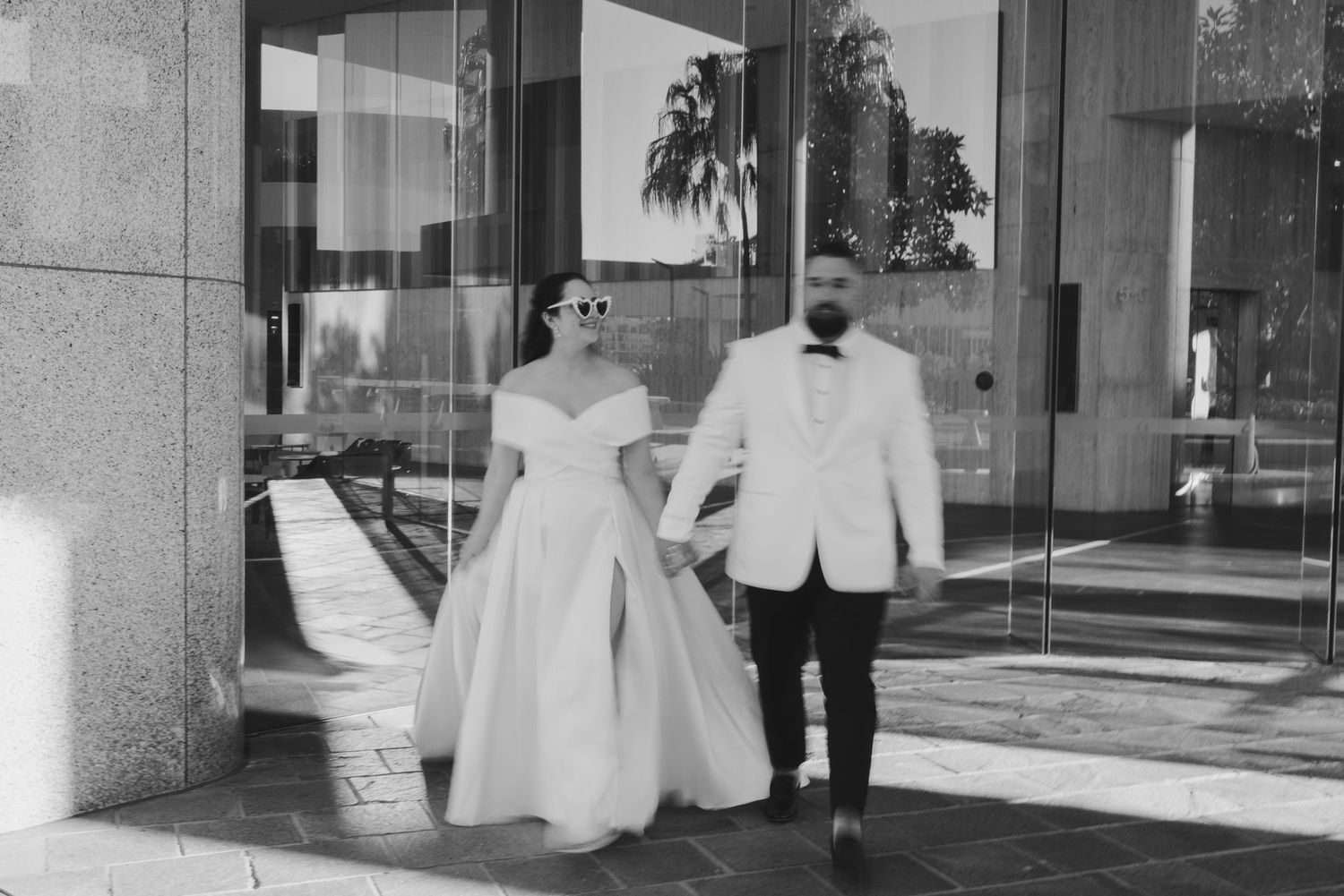 A black and white photograph captures a couple in wedding attire walking together outside a modern building.